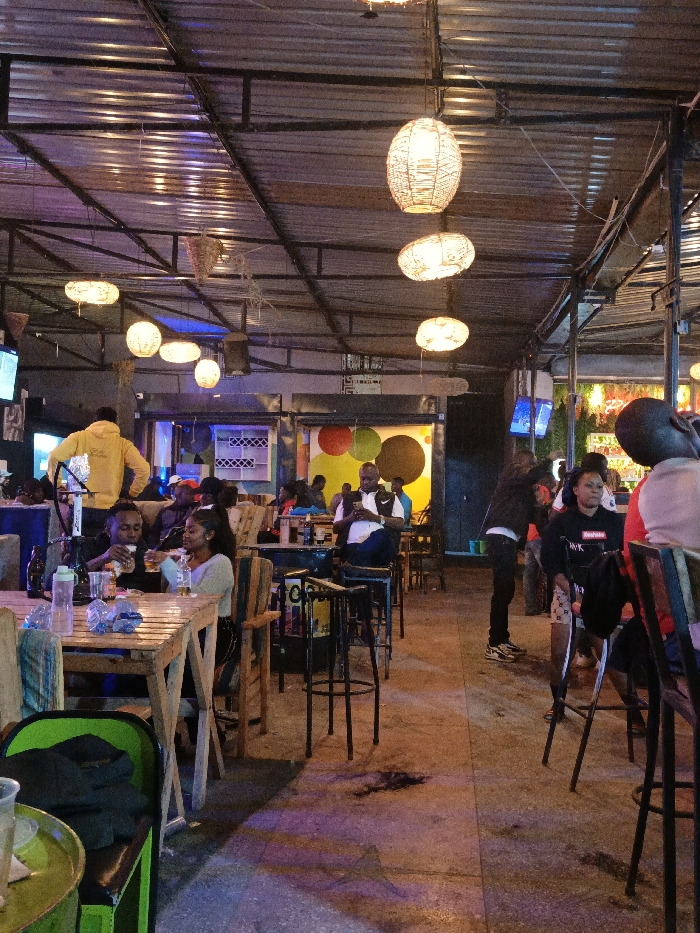 An indoor/outdoor bar area is bustling with patrons. People sit at wooden tables, drinks and personal items scattered about. Others sit at the bar. Overhead, a metal roof structure with hanging wicker lanterns illuminates the scene. The ambiance is casual and social. Several televisions are placed in the background.