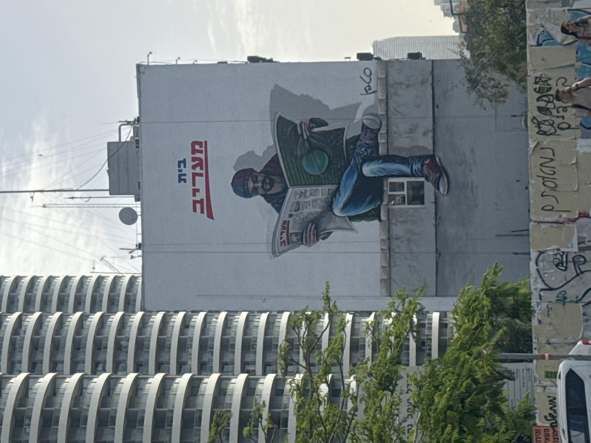The image shows a mural on the side of a building in Tel Aviv, Israel. The mural depicts a man wearing a hat and reading a newspaper with the text "בית מעריב" written in Hebrew. He is sitting with one leg up and wearing jeans and red sneakers. The mural is on a tall, white building with a modern, curved-balcony design in the background. Graffiti covers the corner of the building. Trees and a vehicle are visible in the foreground. The sky is overcast.