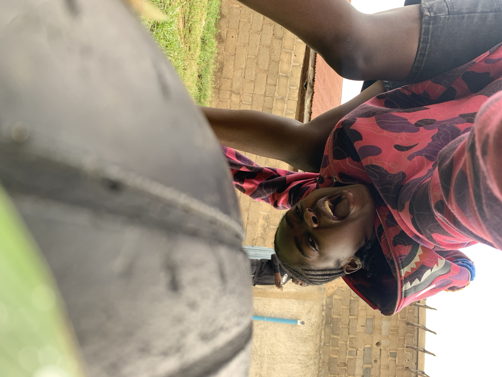The image captures an upward-facing perspective in Rafin Par, Nigeria, showing a person with dark skin and braided hair, wearing a red, black, and white camouflage hoodie and dark shorts, inverted and looking down into the camera with an open mouth. Their legs are visible in the upper left. A large, dark grey, blurry object, consistent with a tire, occupies the bottom foreground. In the background, a light brown brick wall spans the width of the scene, topped with fence spikes on the right. A red corrugated metal roof is visible behind the primary subject's legs. Green grass covers the ground in the mid-left. A second person, dressed in dark attire, stands in the mid-ground to the right of the primary subject, appearing to interact with a mobile phone. A vertical blue pipe or post is visible against the brick wall on the far right. The sky overhead is bright and overcast.
