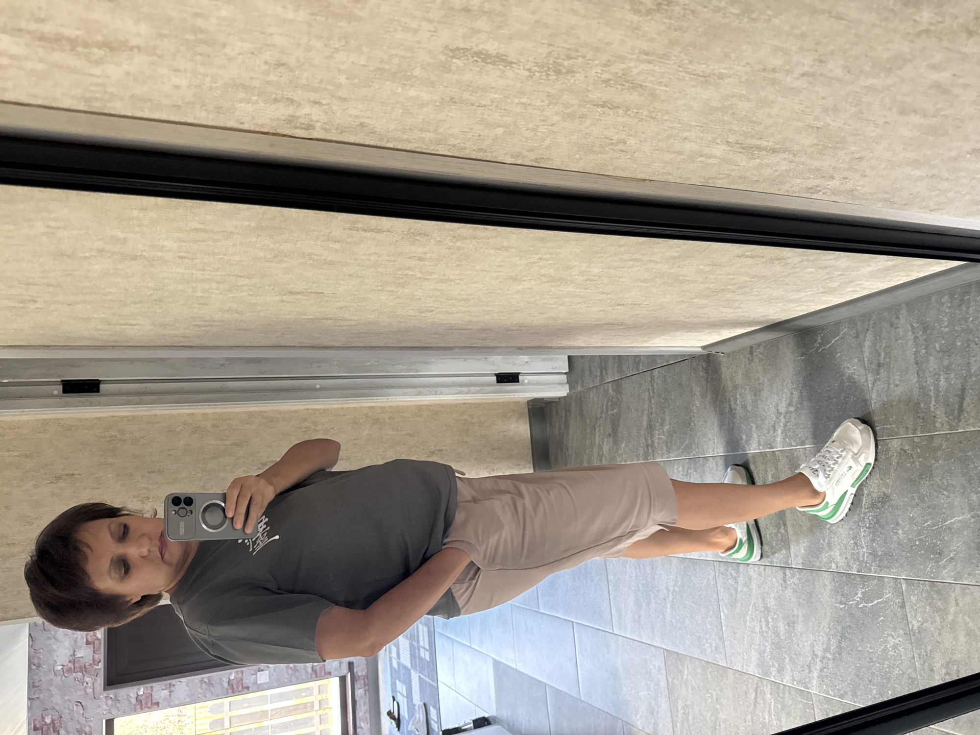 The image shows a woman standing in a room, likely a house, taking a mirror selfie. She is wearing a grey t-shirt and light pink shorts. She has her right hand in her pocket. She is wearing white sneakers with green accents. The room has tiled floors and beige textured walls. In the background, a door and a window with blinds are visible. The woman's hair is dark and styled in a bob. The reflection shows a door and a brick wall with a window. The overall scene suggests a casual indoor setting.