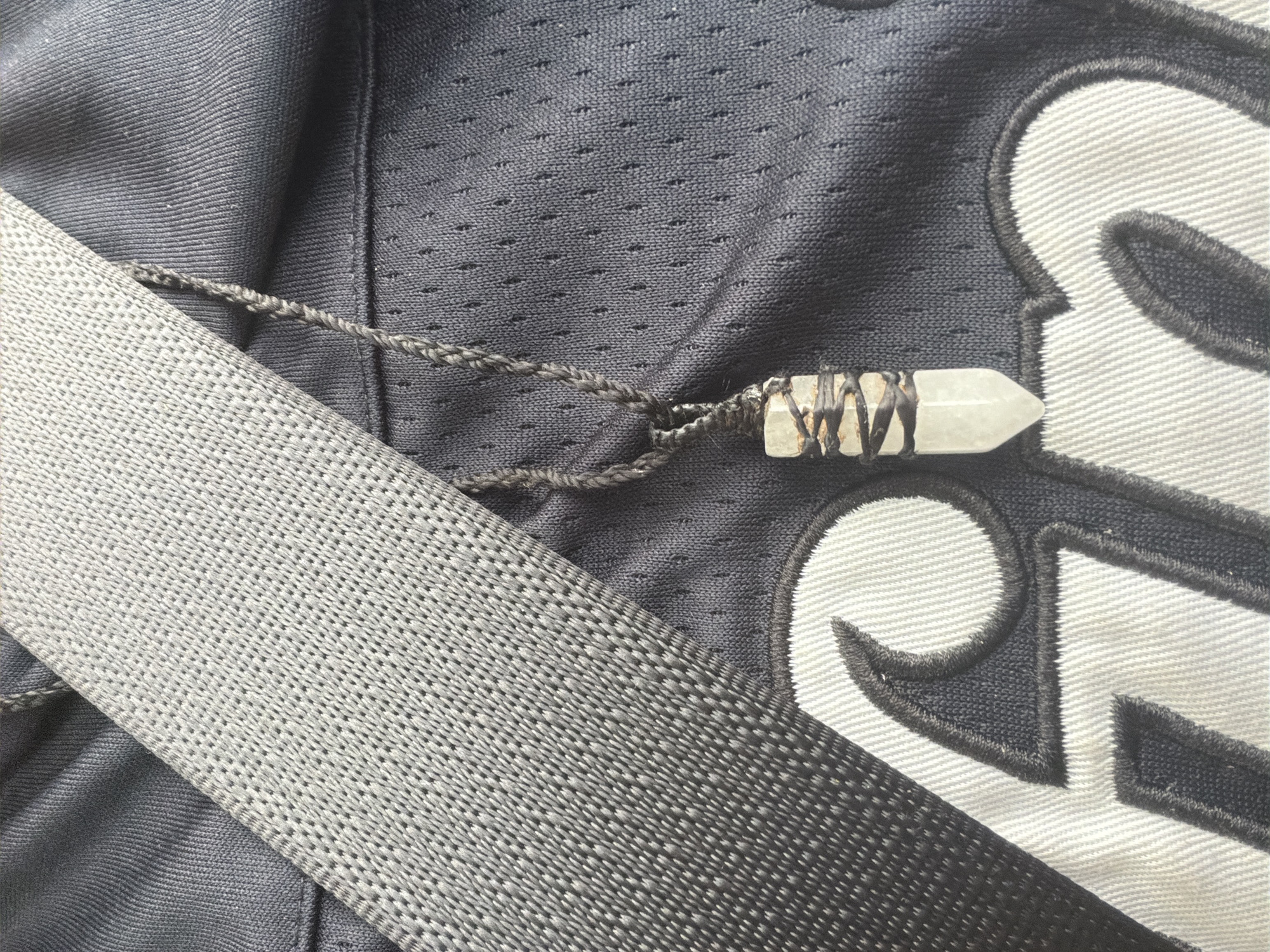 The image shows a close-up of a necklace with a crystal pendant resting on dark fabric with white lettering. A grey, textured strap is diagonally positioned across the upper left portion of the frame. The fabric beneath the necklace appears to be part of a garment, possibly a jersey, due to the patterned texture and the prominent white lettering, which may be an initial or logo. The crystal pendant is elongated and multifaceted, wrapped with dark wire. The scene is devoid of any recognizable urban or natural features that would indicate a specific location in Vallensbæk Landsby, Denmark. The lighting suggests an indoor setting or an overcast day.