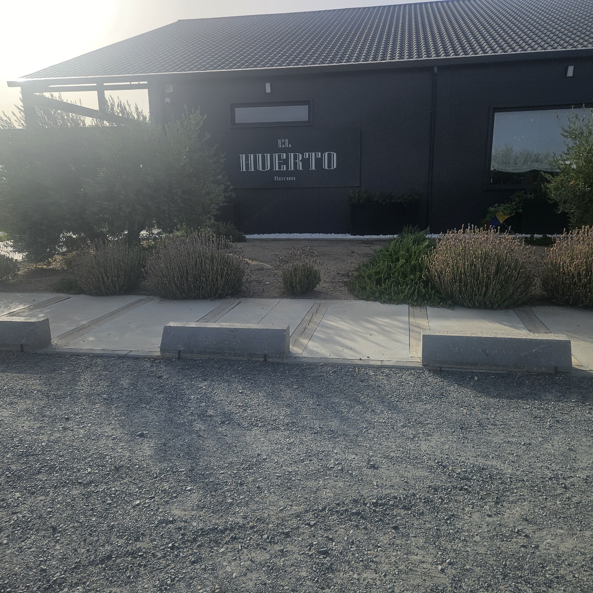 The image shows the exterior of a building with a dark gray facade, likely a business or establishment, bearing the name "EL HUERTO IBERUM" in white lettering. The building has a dark tiled roof and a large window on the right side. In front of the building, there is a gravel-covered area with concrete parking spaces marked by low concrete barriers. To the left of the building, there is a covered walkway or patio area with wooden beams. The foreground is dominated by the gravel and concrete parking area, with some greenery and bushes visible around the building and in the background. The lighting suggests it is daytime, possibly late afternoon or early morning due to the angle of the sun and the shadows. The scene appears to be located in a relatively open area, possibly a rural or semi-rural setting on the outskirts of Illescas, Spain, given the "Iberum" reference which often relates to the Iberian Peninsula. There are no people visible in the image.