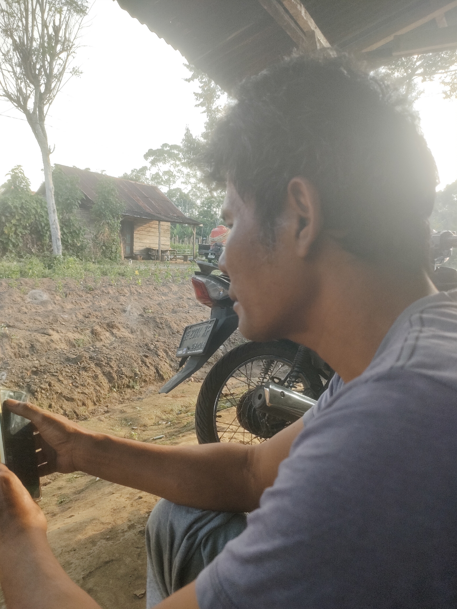 A person is holding a smartphone in their left hand, appearing to be taking a selfie or recording a video, in an outdoor rural setting. The person, facing right in profile, has dark, curly hair and is wearing a purple t-shirt. The background features a partially built structure with a corrugated metal roof, a dirt path, vegetation, and a motorcycle with a license plate. The overall lighting suggests it is daytime, possibly late afternoon or early morning due to the soft sunlight. This scene is located in Padang Dalom, Indonesia.