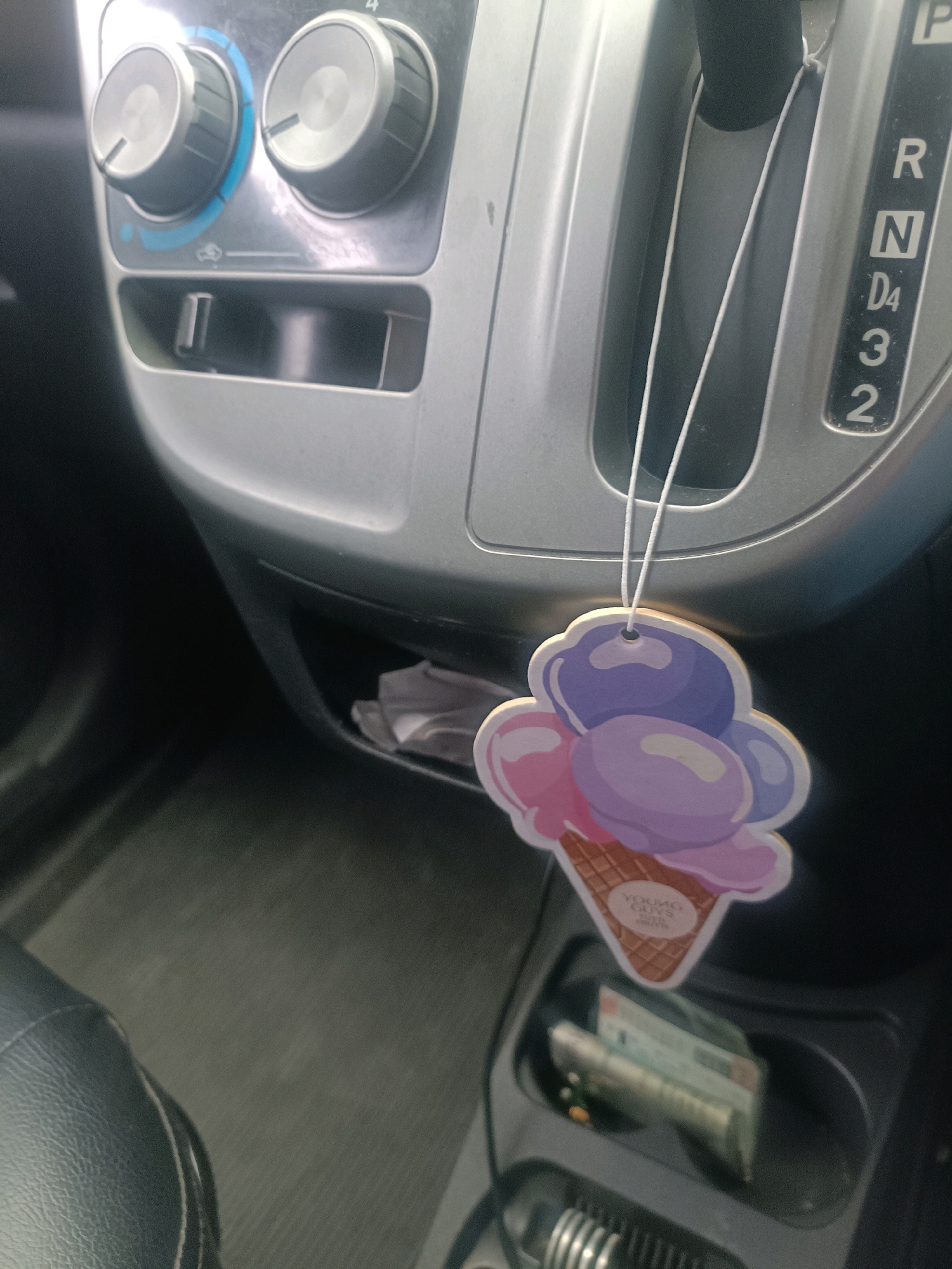 The image shows the interior of a car, focusing on the center console. A colorful ice cream cone-shaped air freshener hangs from the gear shifter. The gear shift is in the "P" (Park) position. To the left of the gear shifter are climate control knobs and a small storage compartment with a USB port. In the lower right of the image, a cup holder contains some folded bills and what appears to be a card. The car's interior is a neutral gray color. The image appears to be taken during daylight, likely inside a parked vehicle. The specific location in Palopo, Indonesia, cannot be determined from the visual information provided.