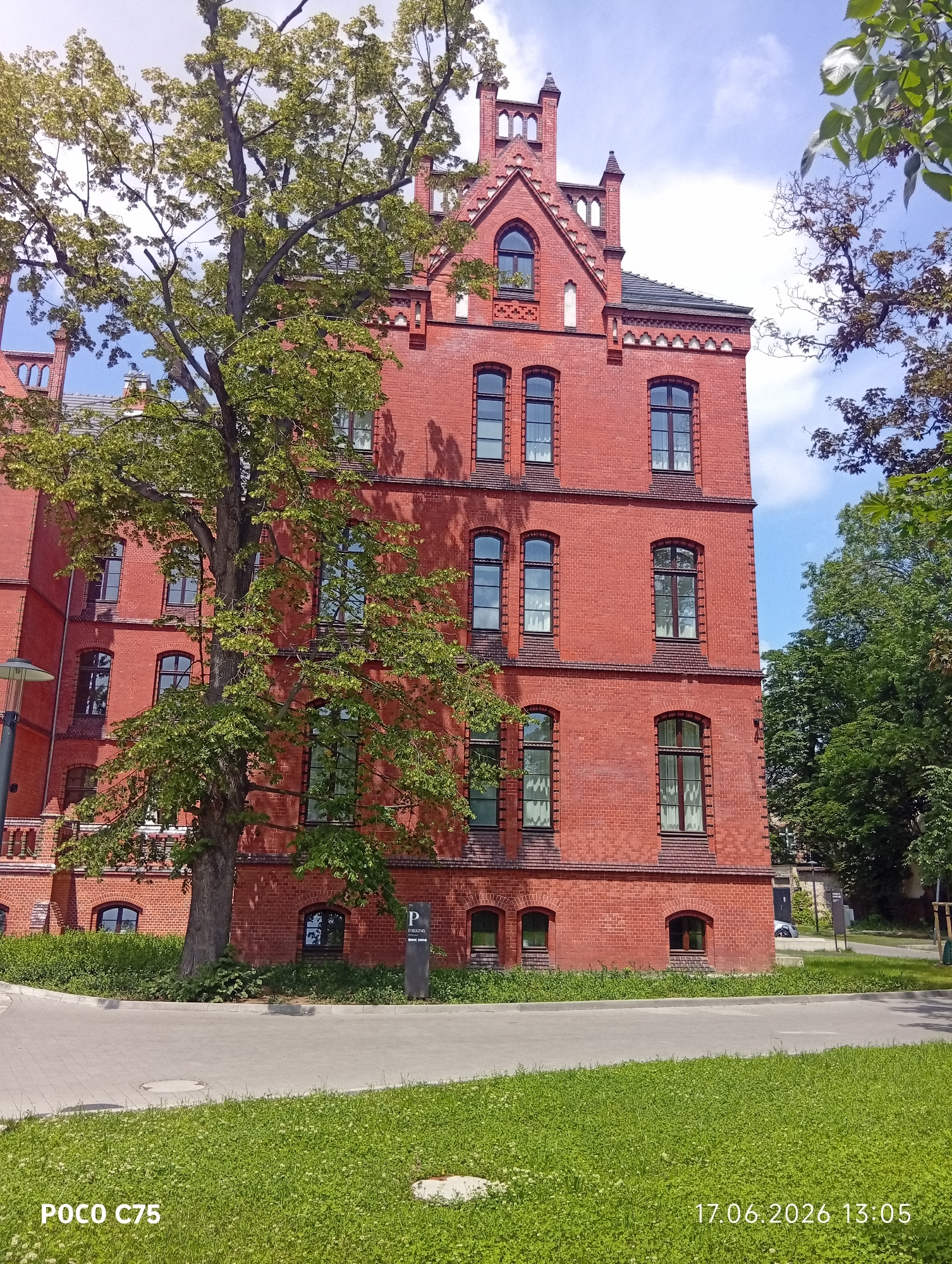 A brick building, likely an older institutional or residential structure, stands tall against a bright blue sky with scattered white clouds. The building is constructed from red bricks and features a steeply pitched roof with a dark shingle covering. It has multiple floors with numerous windows, many of which are arched, some with decorative detailing above them. A large, leafy tree with a thick trunk partially obscures the left side of the building, its branches reaching towards the structure.
In the foreground, a paved pathway curves around a well-maintained lawn. The grass is lush and green, dotted with small white flowers. To the right of the pathway, a dark sign with a white "P" and the word "PARKING" indicates a parking area. Beyond the building, more trees and foliage suggest a park-like or campus setting. The scene appears to be bathed in bright sunlight, indicating it is daytime.
Visible text includes "POCO C75" in the bottom left corner and "17.06.2026 13:05" in the bottom right corner, suggesting the image was taken on June 17, 2026, at 1:05 PM. The overall impression is one of a pleasant, sunny day in an urban or suburban environment, with a focus on the architectural details of the building and the surrounding greenery.