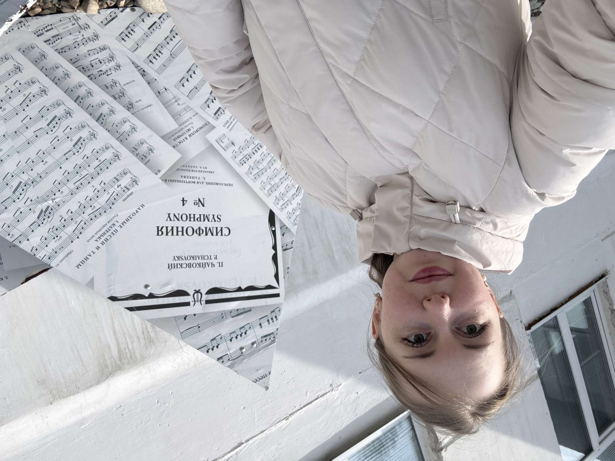 A fair-skinned young woman with light brown hair stands in an outdoor environment, possibly in the city of Unknown, Unknown. She wears a beige quilted jacket and is positioned next to sheet music, with a cover indicating Tchaikovsky's Symphony No. 4 in Russian. Additional sheets display musical notation. A white wall and a window with white frames are visible in the background, suggesting an exterior setting, perhaps near a building. The image is rotated, causing the subject to appear upside down.