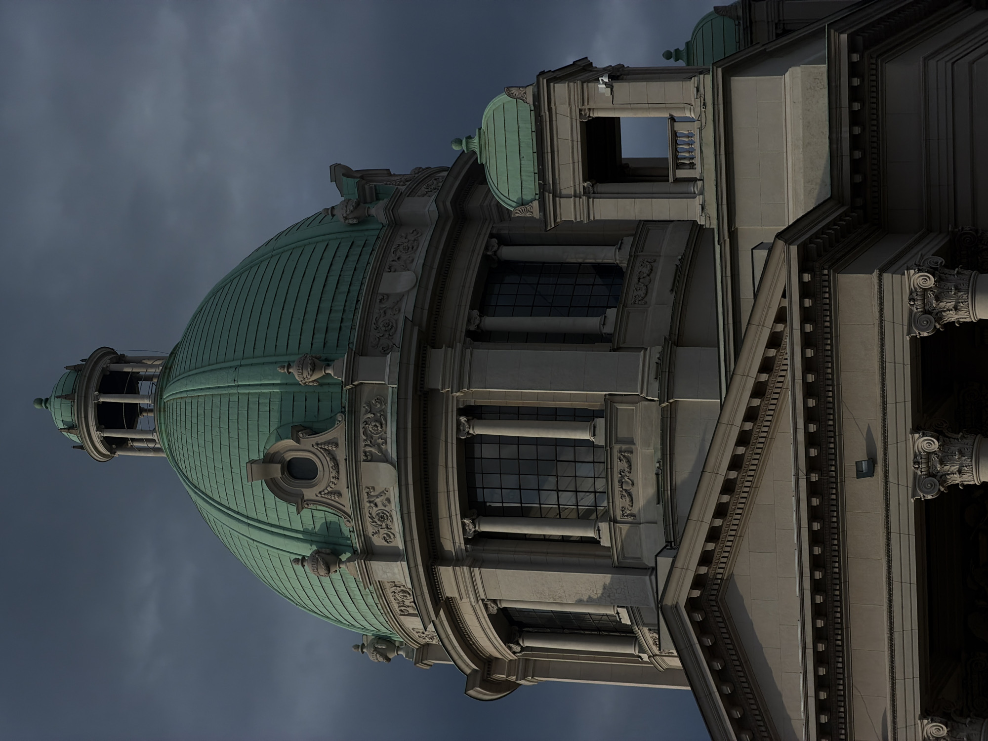 The image shows a close-up view of the dome and upper facade of the National Assembly of Serbia building in Belgrade, Serbia. The building is constructed of light-colored stone and features classical architectural elements, including columns, ornate carvings, and a prominent green copper dome. The dome is segmented and topped with a smaller cupola. The sky behind the building is dark and cloudy, suggesting a dramatic or overcast day. The lighting emphasizes the architectural details and creates strong shadows. There are no people visible in the image.
