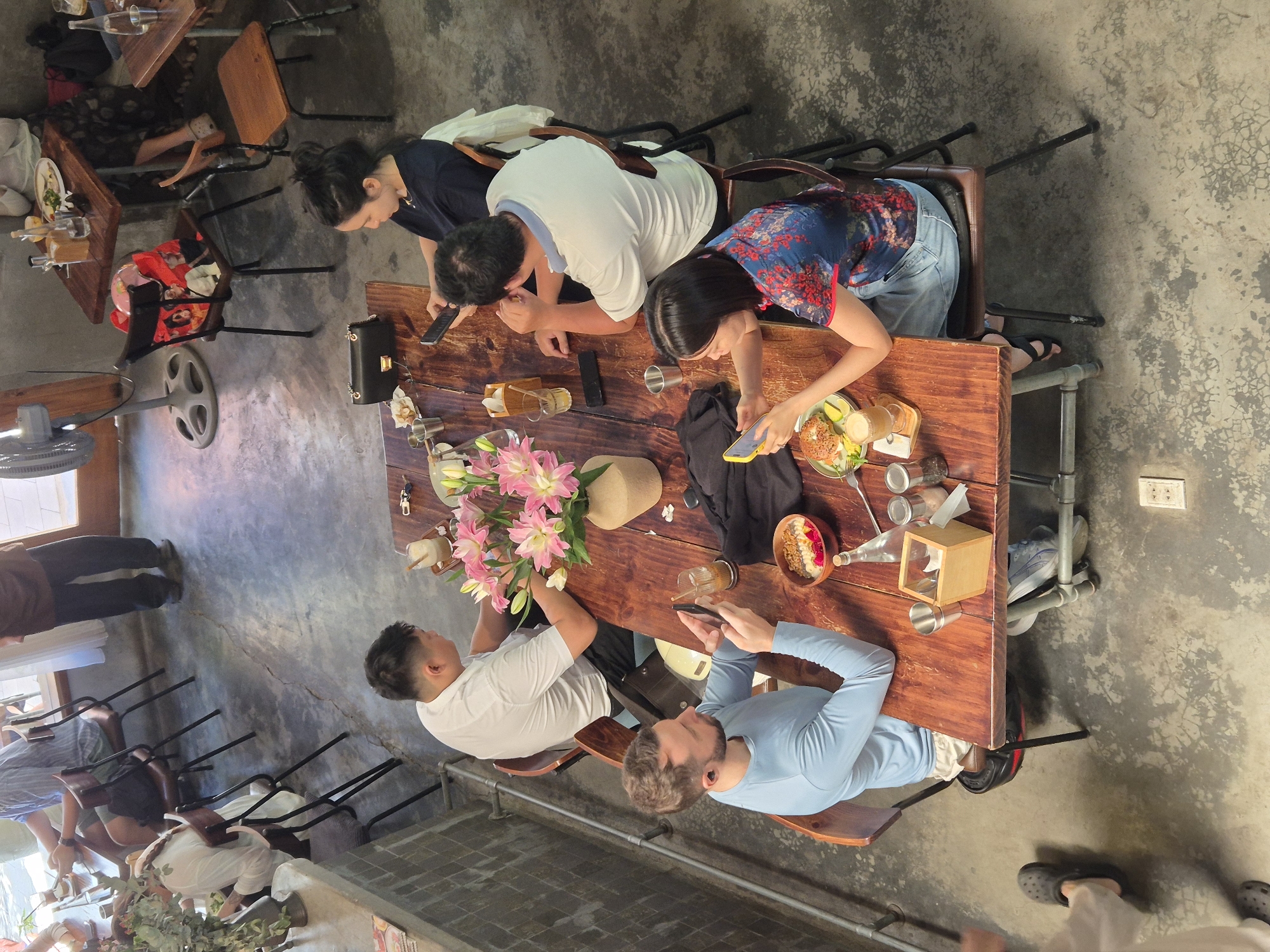 The image depicts an overhead view of a group of people seated around a long, rustic wooden table in what appears to be a cafe or restaurant in Da Nang, Vietnam. The floor is a mottled grey concrete. On the table, there are various food items, drinks, and personal belongings. A large vase of pink lilies is placed centrally on the table. Several individuals are engaged with their mobile phones. One person in a light blue long-sleeved shirt is looking at their phone, holding it in both hands. Another person in a blue and red patterned shirt is also looking at their phone. Further up the table, a person in a white shirt is looking down at their phone, and a person in a dark blue shirt is also focused on their phone. Two individuals are partially visible at the far end of the table. On chairs around the table, there are bags and other items. A large industrial fan is visible in the background, and some patrons are seated at other tables in the distance. The lighting suggests it is daytime, possibly with natural light filtering in from outside.