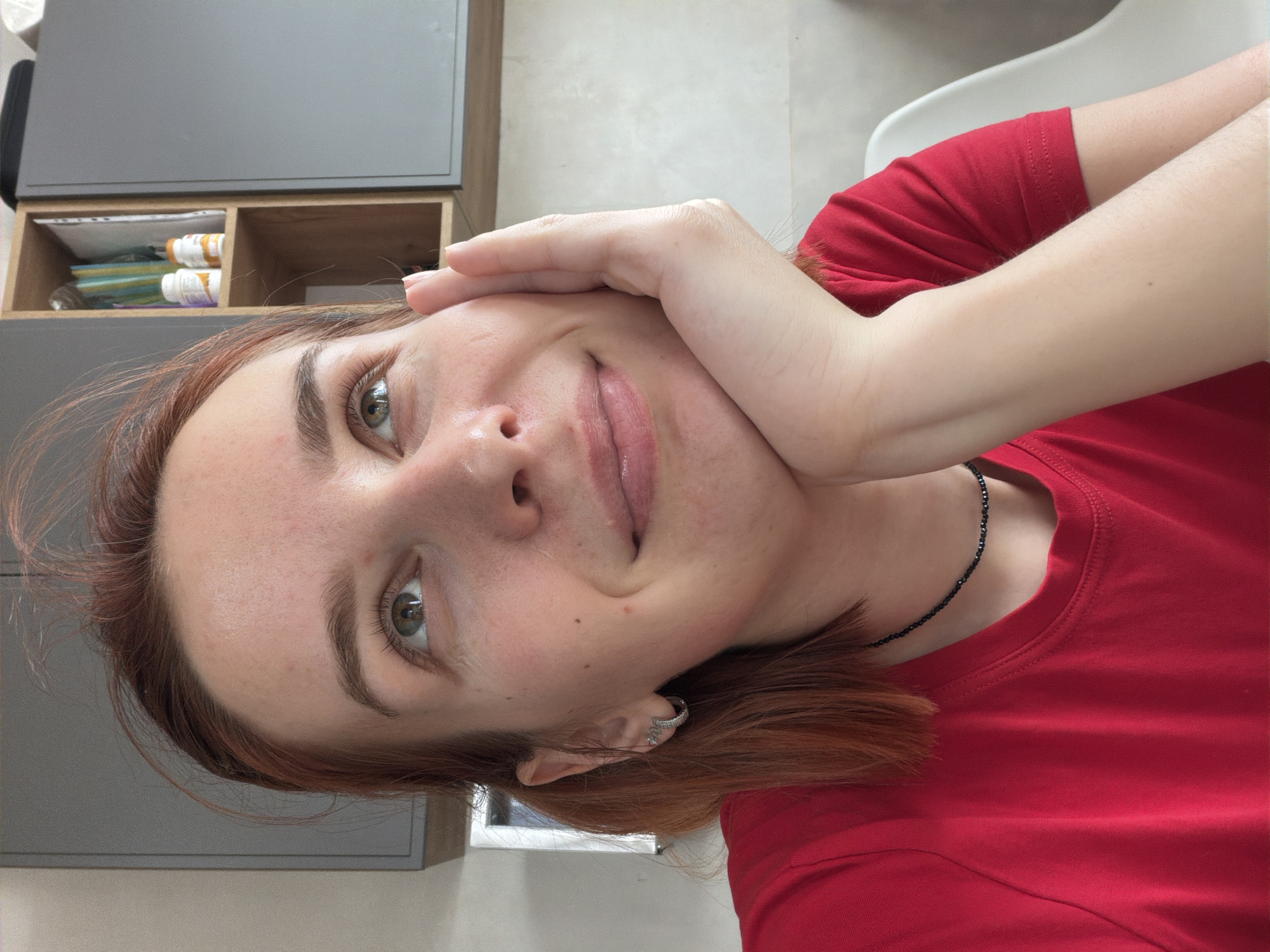 The scene is an indoor setting in Chaikovsky, Russia, depicting an adult female from the chest up. She has fair skin, reddish-brown hair, and green-hazel eyes, with a slight smile on her face. She is wearing a bright red short-sleeved t-shirt, small hoop earrings in her left ear, and a black beaded choker necklace. Her right hand is positioned against her jawline, supporting her head, while her gaze is directed upwards and to the viewer's right. The background features light-colored walls and furniture. A grey wall-mounted cabinet is visible on the left. To the right, a wooden open shelving unit is present, containing documents or books on the top shelf, and two white plastic bottles with orange caps on the middle shelf. Adjacent to the wooden unit is another grey wall-mounted cabinet. A portion of a white chair back is visible in the lower right of the frame.