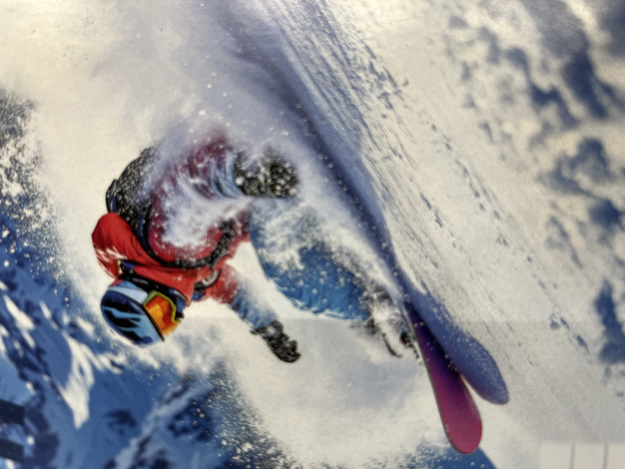 The image shows a snowboarder in action, carving down a snowy slope and creating a spray of snow. The snowboarder is wearing a red jacket, blue pants, a helmet, and goggles. The background consists of snowy mountains. The image is a photograph and does not depict any location within the city of Kharkiv, Ukraine.