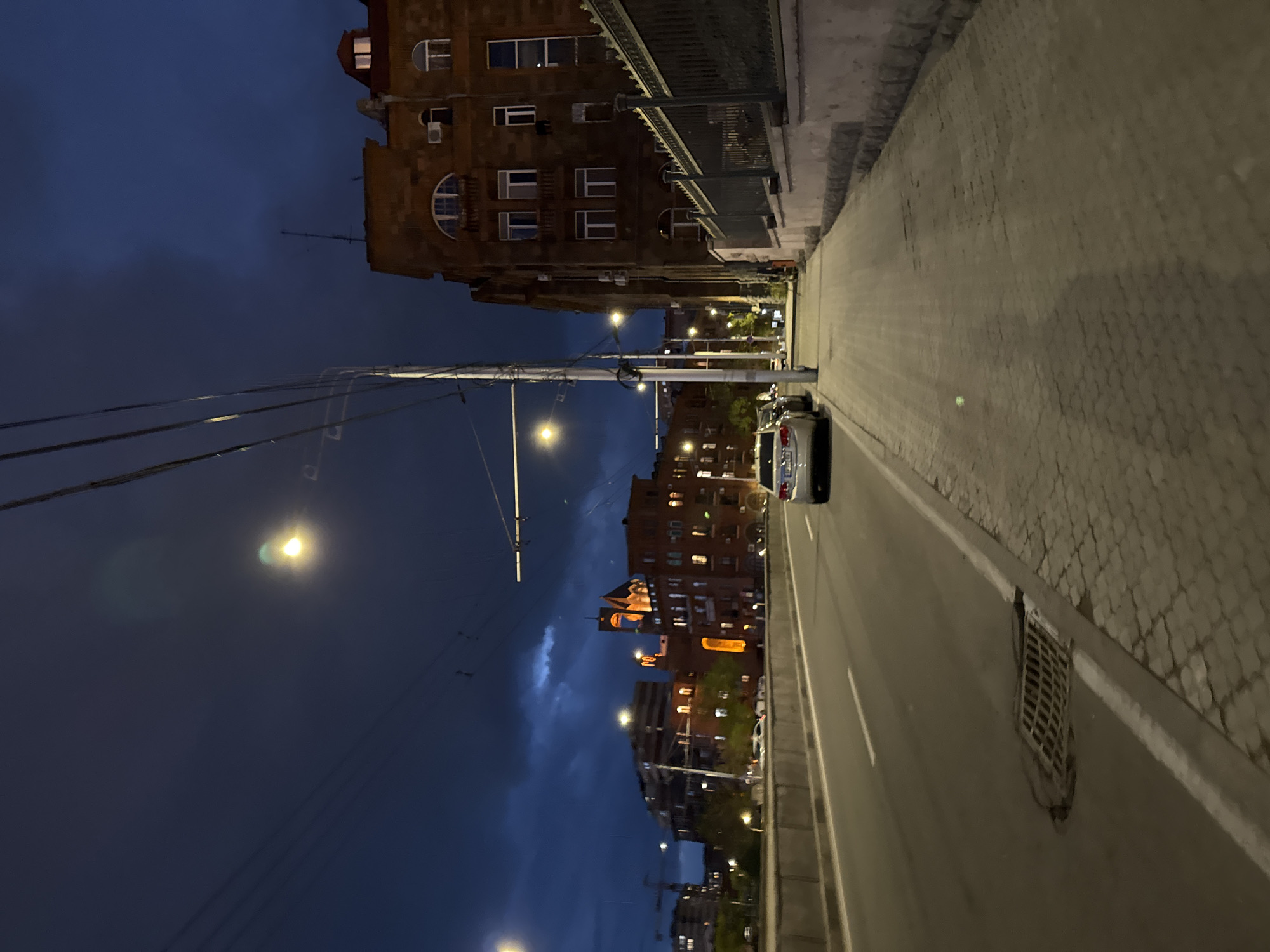 This image presents an urban street scene in Yerevan, Armenia, captured at night or late dusk from a slightly elevated perspective. The dark blue sky, partially obscured by subtle cloud formations, is punctuated by bright streetlights casting warm, yellow-orange glows and noticeable lens flares.
On the left side of the frame, a dense network of utility poles and overhead wires is prominent, suggesting a bustling urban infrastructure. Beyond these, multi-story buildings constructed of reddish-brown tuff stone or brick line the street. Many windows within these buildings are illuminated, showcasing a characteristic architectural style that includes both rectangular and arched windows, along with visible air conditioning units.
The road, made of asphalt, stretches into the distance, marked by white lane lines. To the right, a wide sidewalk, appearing to be paved with cobblestones or pavers, runs parallel to the road. A silver or white car is visible on the right side of the road, with other vehicle lights discernable further down the street. A metal drainage grate is embedded into the sidewalk's edge in the foreground. No human activity or discernible text is visible, conveying a peaceful, quiet urban atmosphere.