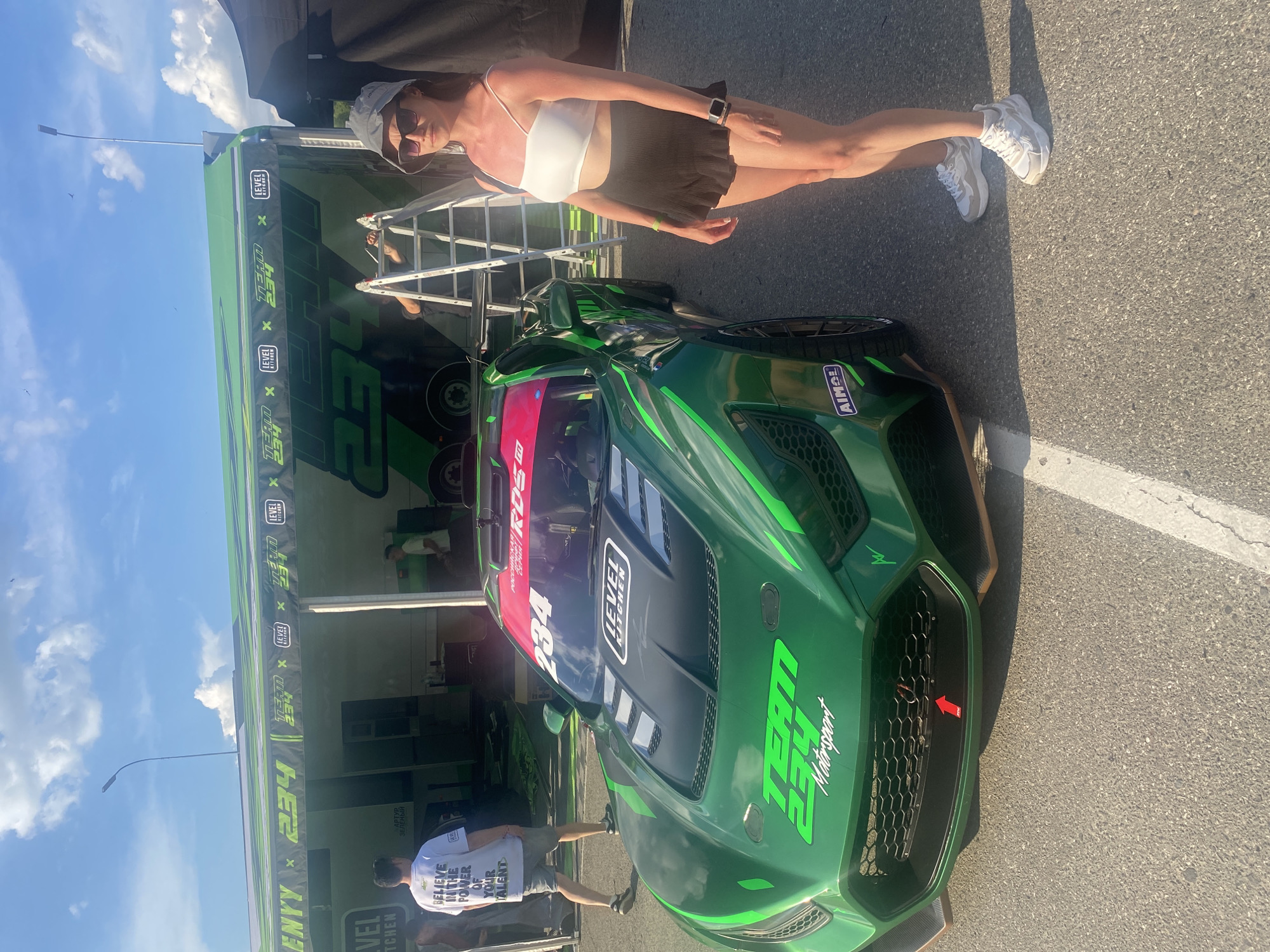 A green, low-profile sports car with a black hood and racing decals is parked on asphalt. The car is adorned with the logos "TEAM 234 Motorsport," "LEVEL KITCHEN," and a red tow hook with an upward arrow sticker. The car's windshield has a white number "234" in a red circle and the word "ROS" on a pink sticker. To the right of the car, a young woman wearing a white crop top, a black pleated skirt, white sneakers, a grey bucket hat, and sunglasses stands facing the camera. Behind the car, a large green and white trailer with "TEAM 234" branding is visible, with a ladder leaning against it. Several individuals are seen in the background, including a man in a white t-shirt with the text "BELIEVE IN THE POWER OF YOUR TALENT" walking away from the camera. The environment appears to be an outdoor event or race track under a blue sky with scattered clouds. The scene is set in the city of Beryozovka, Russia.