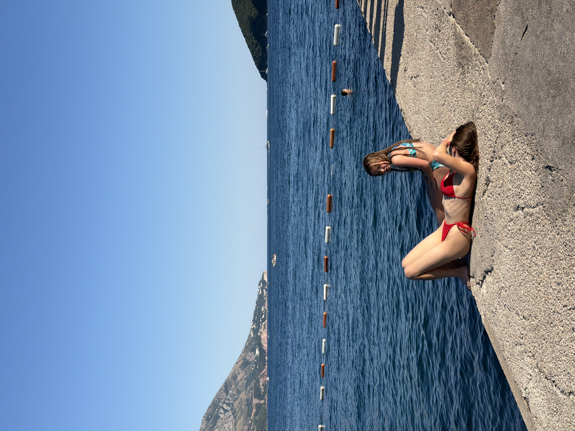 The image shows a bright, sunny day at a waterfront in Budva, Montenegro. The scene is dominated by the deep blue of the Adriatic Sea, bordered by a textured concrete walkway and a rocky, tree-covered mountainside in the distance. A line of orange and white buoys marks a swimming area in the water.
Two young women in bikinis are the primary human subjects. One, wearing a red bikini, is sitting on the edge of the concrete, with her legs bent and her feet dangling over the water. The other, in a blue and green patterned bikini, is standing slightly behind her, leaning forward and looking out towards the sea. Their poses suggest a relaxed and perhaps contemplative moment before swimming or after being in the water.
In the distance, a lone swimmer is visible in the water, and a small boat is sailing on the horizon. The clear blue sky indicates good weather, likely mid-day given the strong sunlight casting shadows on the walkway. The overall atmosphere is one of leisure and enjoyment of a summer day at the coast. No text is visible in the image.