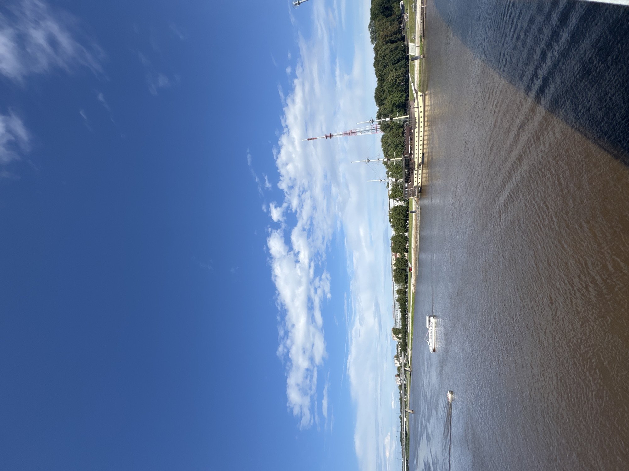 This is a photograph taken from a high vantage point, looking down upon the Volkhov River in Veliky Novgorod, Russia. The sky is a vibrant blue with scattered white clouds.

In the river, two boats are visible. A larger white boat with a canopy is moving away from the viewer towards the left. Further to the left and closer to the viewer, a smaller, red-and-white boat is also moving away.

On the far bank of the river, a long, multi-story building with many windows is situated behind a line of green trees. To the right of this building, a historical wooden ship is moored. The ship has multiple masts and a light-colored hull with dark windows. Behind the ship, a tall, red-and-white telecommunications tower rises into the sky. A bridge is visible in the distance to the left. The water of the river is a dark, muddy brown, reflecting the sky. A dark shadow, cast by the photographer's position, diagonally bisects the lower right portion of the image.