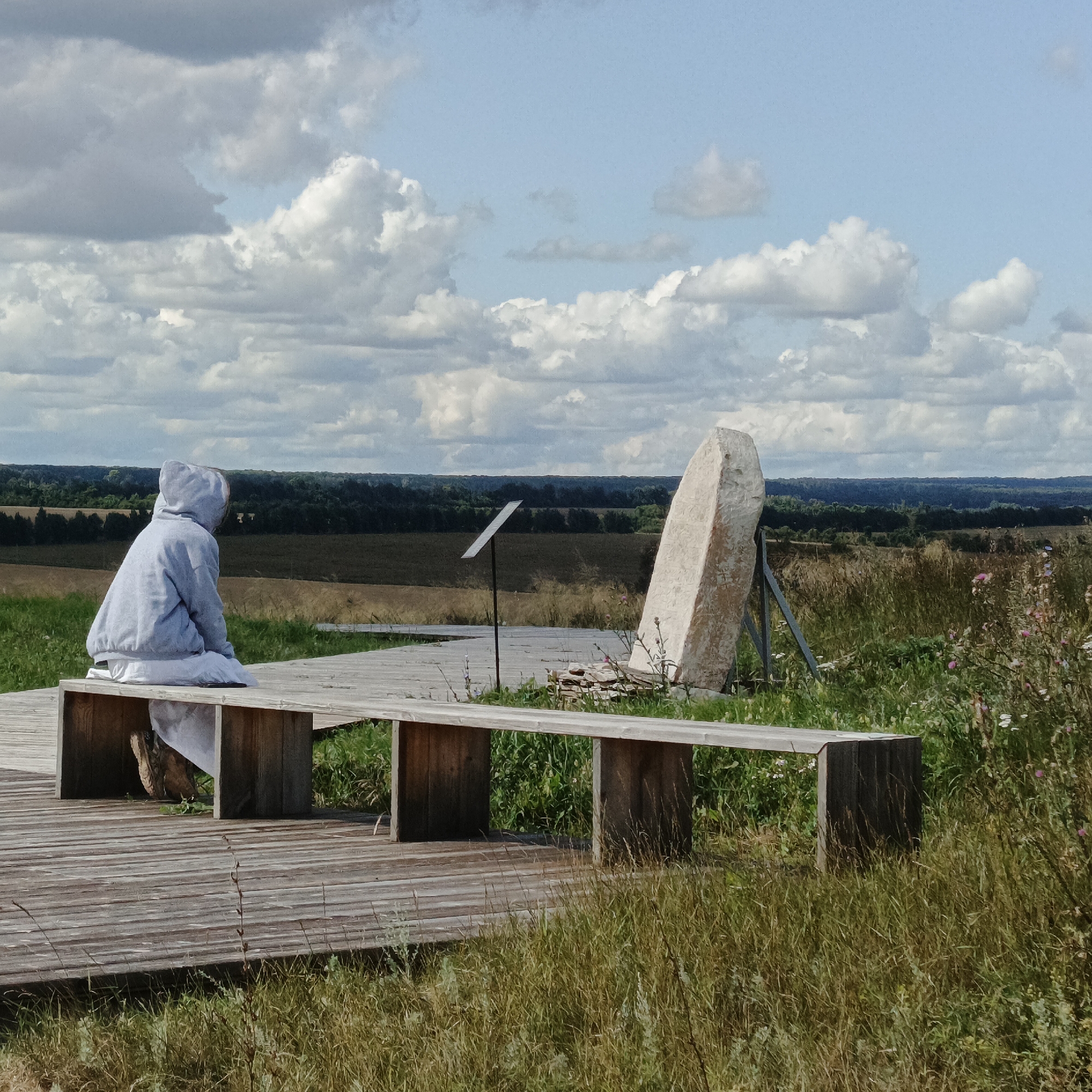 In Verkhnie Termy, Russia, a person wearing a hooded grey jacket and white pants sits on a wooden bench facing away from the camera. The bench is situated on a wooden boardwalk overlooking a valley. To the right of the bench, a large, rough-hewn stone stands upright, supported by metal poles. In the background, a dense line of trees borders a vast, cultivated field under a sky filled with scattered white clouds. The environment is predominantly rural and natural.