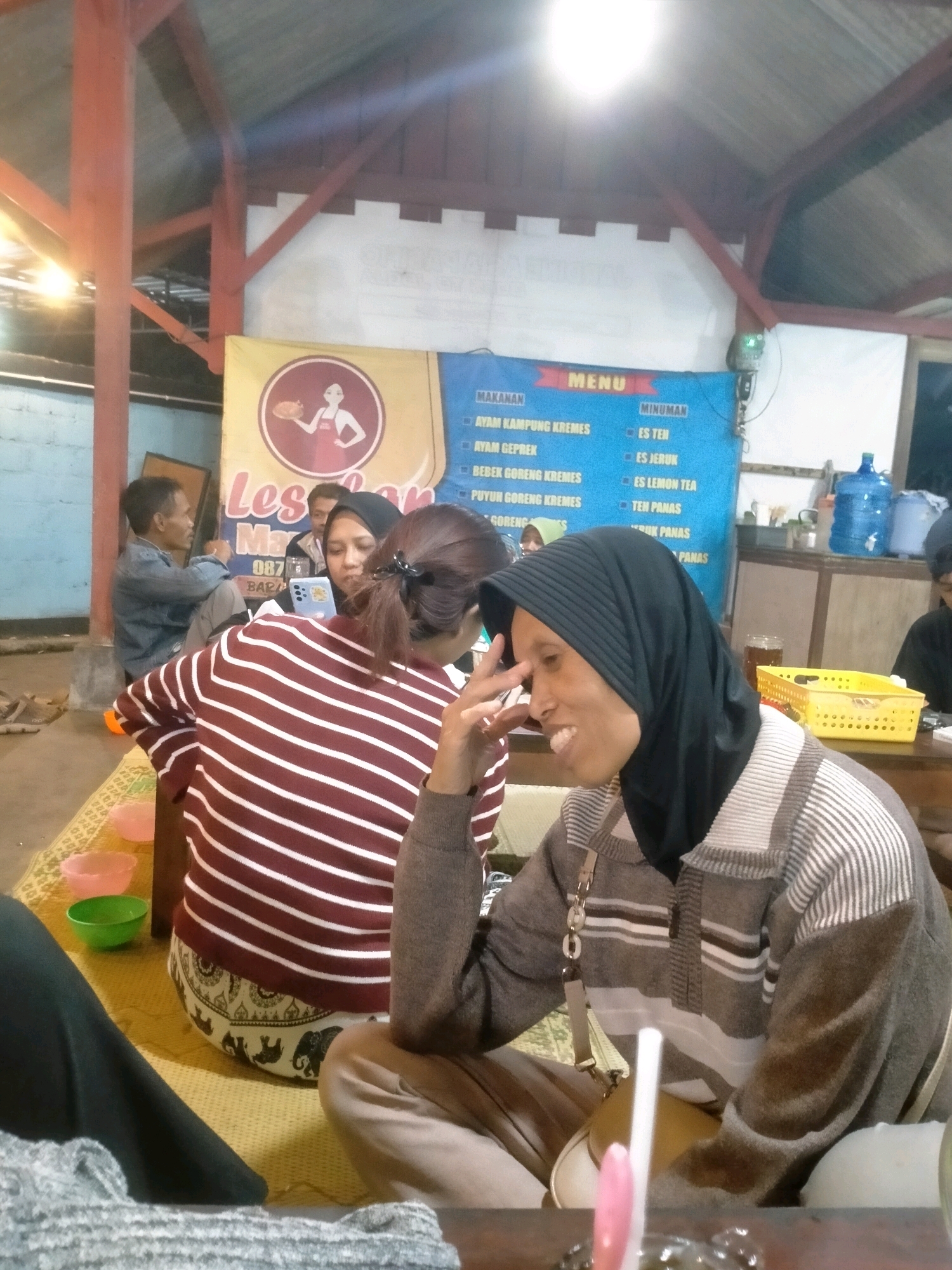 A group of people are gathered around a table at a food stall in Playen, Indonesia. The stall is covered by a corrugated metal roof supported by wooden beams. A banner in the background advertises "Les" and lists food and drink options. A woman in a black hijab and a striped sweater is seated in the foreground, smiling and looking to her right. Another woman in a red and white striped shirt is seated to her left, with her back to the camera. A man in a denim shirt is visible in the background, seated and looking at the people at the table. The atmosphere appears casual and social, with the people engaged in conversation. The presence of food bowls and a water dispenser suggests a dining setting.