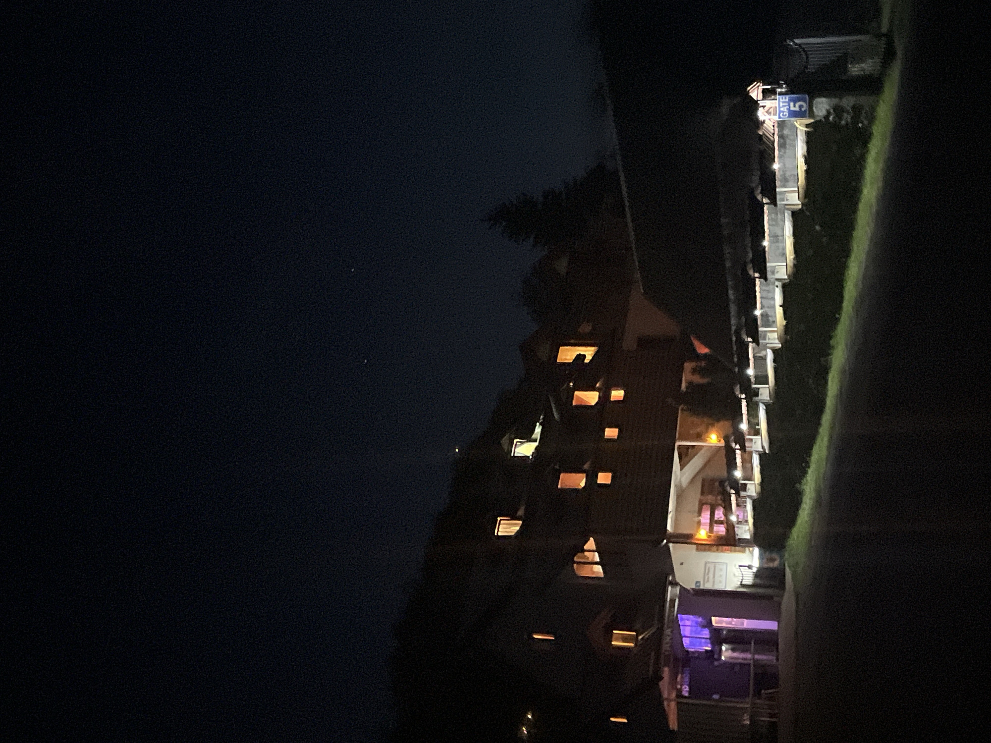 At night, a multi-story building with lit windows dominates the right side of the frame, suggesting it is located in Polja, Montenegro. The lighting indicates activity within the building. Adjacent to the building, there's a sign reading "Gate 5". Adjacent to the gate is a grassy area. The left side is mostly dark, but some lights in the sky are visible.