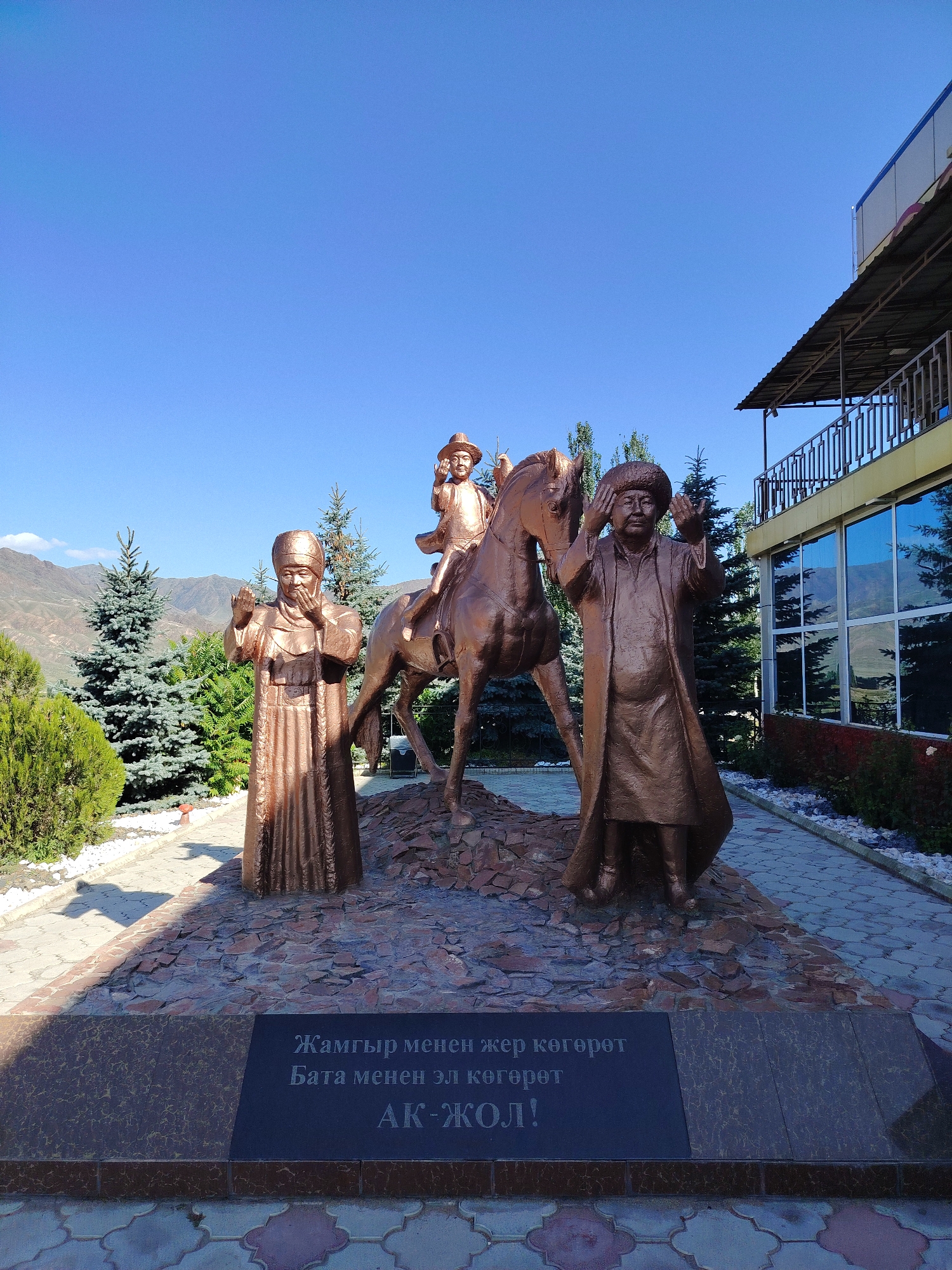 This is a medium shot, eye-level photograph of a bronze sculpture in a city park in an unknown location in Kyrgyzstan. The sculpture depicts three figures: a man on horseback, a man standing to the right, and a woman standing to the left. The man on horseback is wearing a hat and appears to be a child. The standing figures are dressed in traditional clothing. The man on the right is wearing a hat and robe, with his hands raised. The woman on the left is wearing a headscarf and robe, with her hands also raised. The sculpture is set on a raised stone base. In front of the sculpture is a dark stone plaque with text inscribed in Cyrillic script. The background features evergreen trees, mountains, a clear blue sky, and a modern building with large windows and an overhang. The ground in front of the sculpture is paved with decorative tiles. The lighting suggests it is daytime, with shadows cast by the figures.