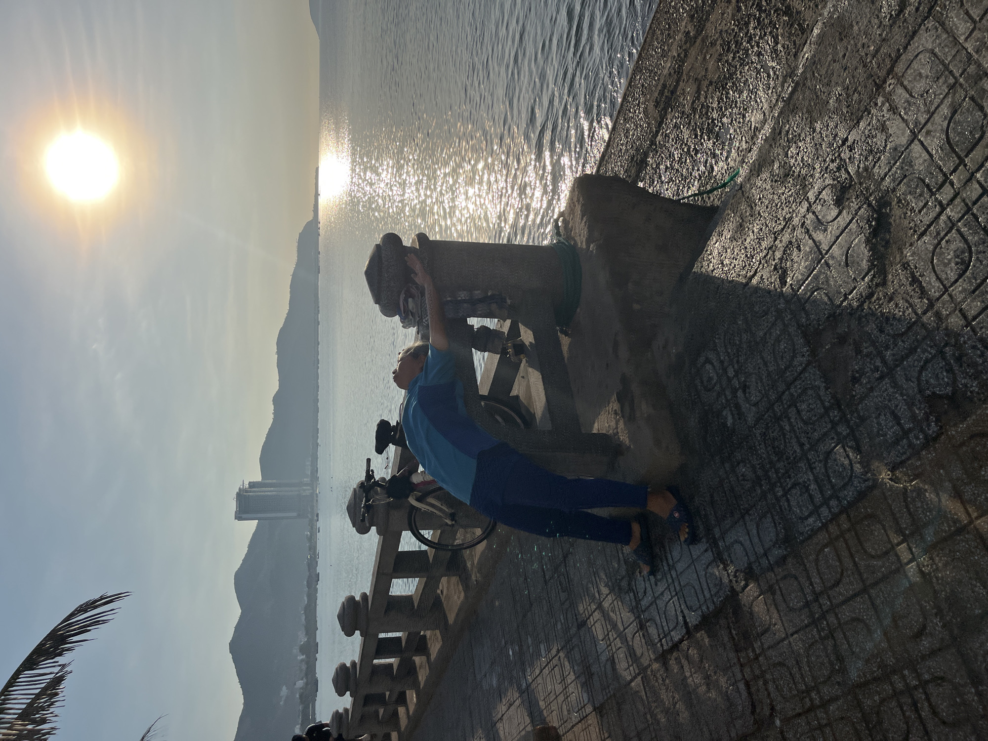 A person in a blue shirt and pants is leaning against a concrete cannon at the water's edge in Bac Nha Trang Ward, Vietnam. The person has their hands on the top of the cannon. A bicycle is parked nearby on a tiled walkway along a low concrete wall. In the background, the sun is reflecting on the water, with mountains and buildings visible on the horizon.