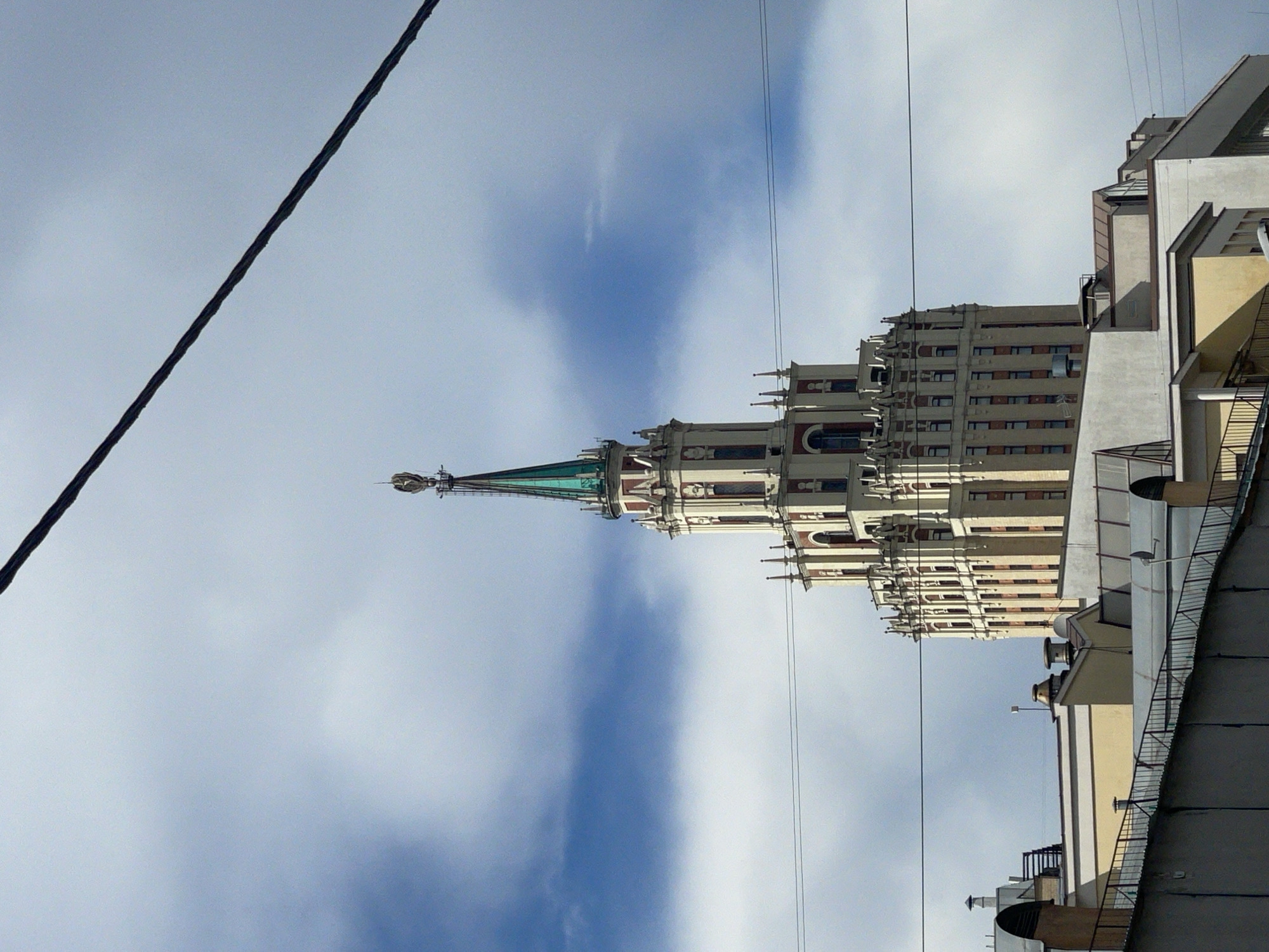 The image shows a view of a tall, ornate building with a spire, characteristic of Stalinist architecture, against a cloudy sky. Several other buildings, including a modern apartment complex, are also visible. Wires are strung across the sky, a common sight in urban environments. The perspective is from a low angle, looking up at the structures. The lighting suggests it is daytime, likely midday, with diffused sunlight from behind the clouds. The overall scene conveys a sense of a typical urban landscape in Moscow, Russia, characterized by a mix of historical and modern architecture. No people are visible in the frame. There is no visible text.