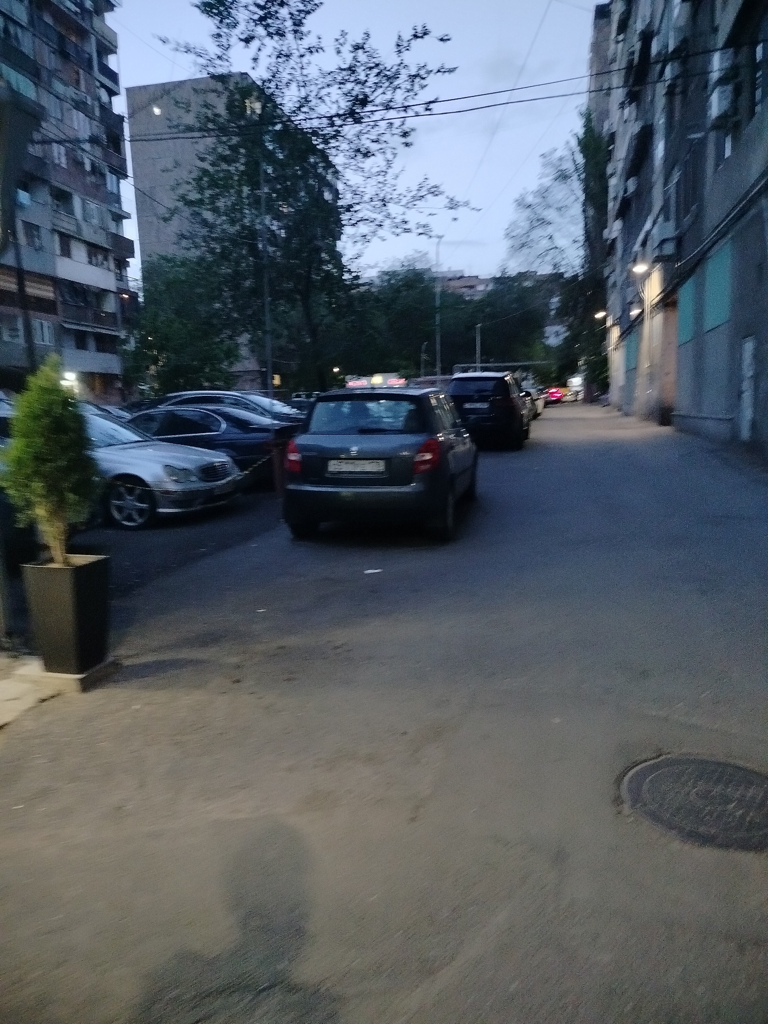 The image depicts an urban street scene in Yerevan, Armenia, at dusk. The street is lined with tall, Soviet-era apartment buildings on both sides. Cars are parked along the left side of the street, with a dark-colored Škoda Fabia prominently in the center, viewed from the rear. A small, potted evergreen tree stands in the lower left corner. In the distance, more cars are visible, suggesting a residential area. The street surface appears worn, and a manhole cover is visible in the lower right corner. The sky is overcast.