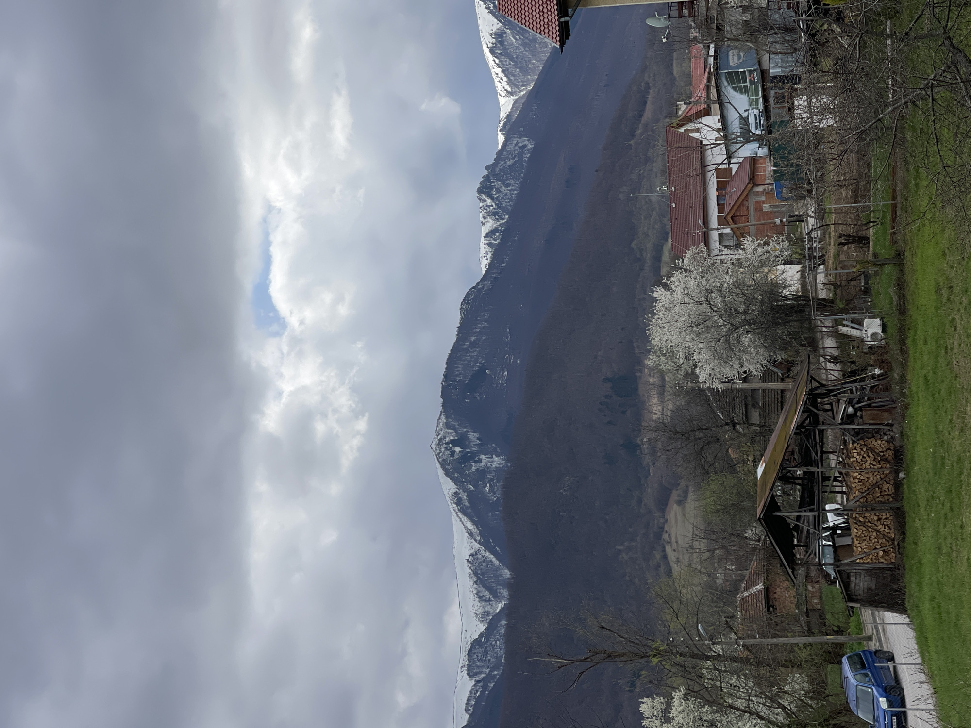 The image shows a view in the city of Varshets, Bulgaria. The foreground features a residential area with houses, trees in bloom, and a stack of firewood under a shed. A blue car is parked nearby. In the background, a mountain range with patches of snow is visible under a cloudy sky.