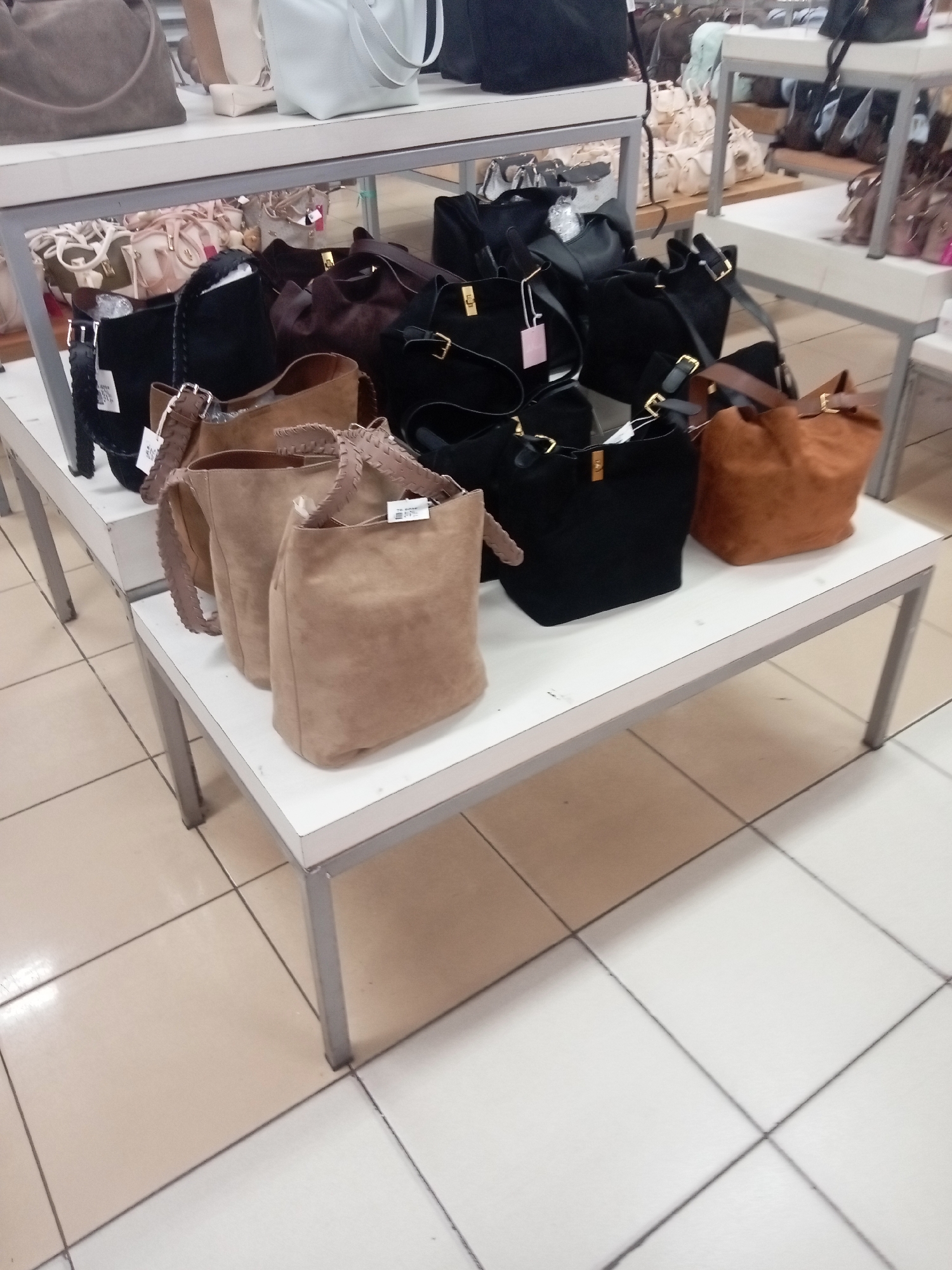 The image shows a retail display of handbags on white shelves in a store. The handbags are of various colors and materials, including suede and leather. Some of the handbags are black, brown, and tan. The display is arranged in multiple rows on the shelves. The background appears to be a retail environment with tiled flooring. The overall scene is static and does not depict any people or ongoing actions. The location is identified as Batam, Indonesia.