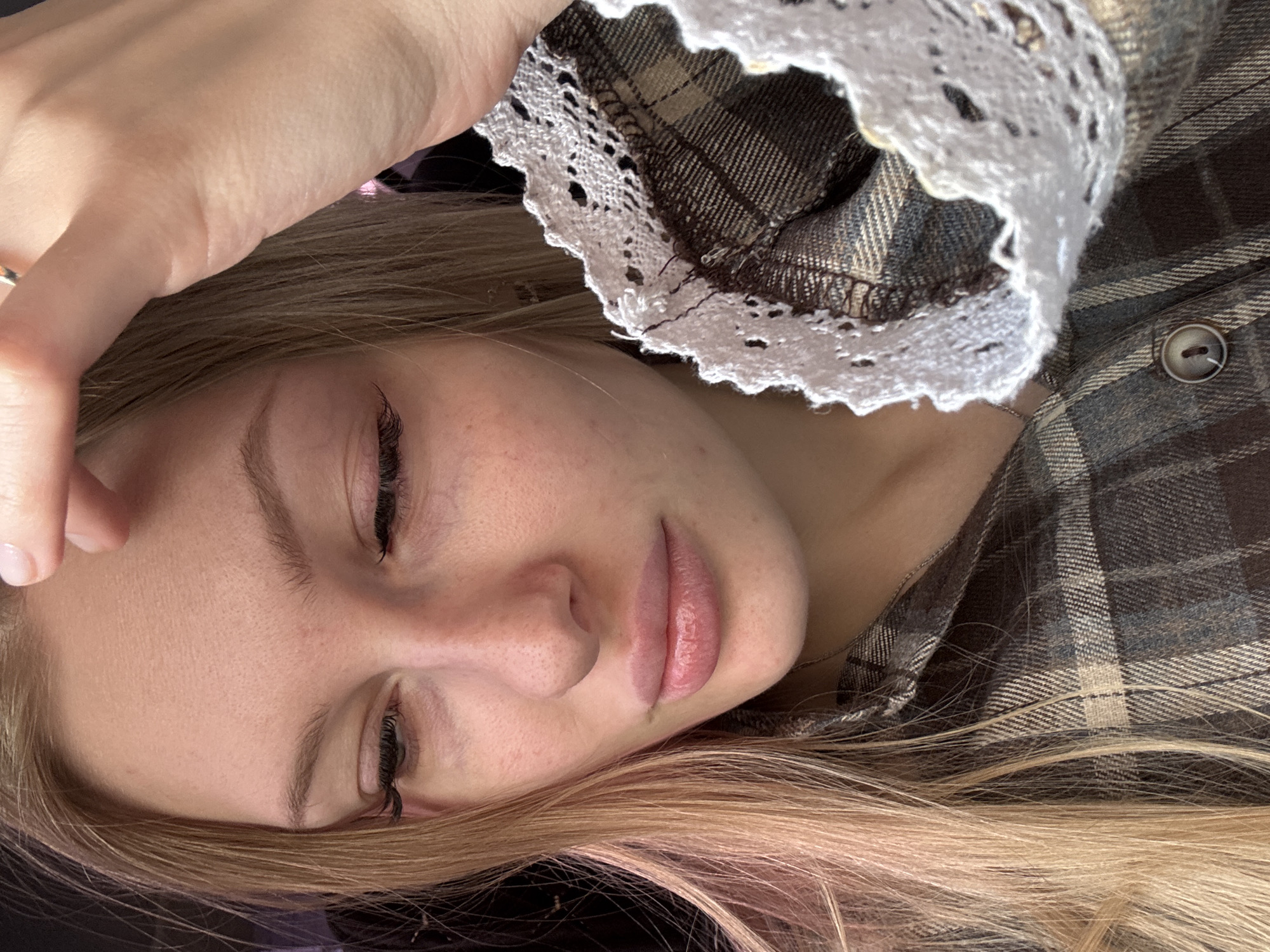 The image shows a person with fair skin, light blonde hair with some pink highlights, and closed eyes. The person's right hand is touching their forehead. The person is wearing a brown and grey plaid shirt with a white lace collar. A button is visible on the shirt. The location is not discernable from the image, therefore a city in Russia is not deducible.