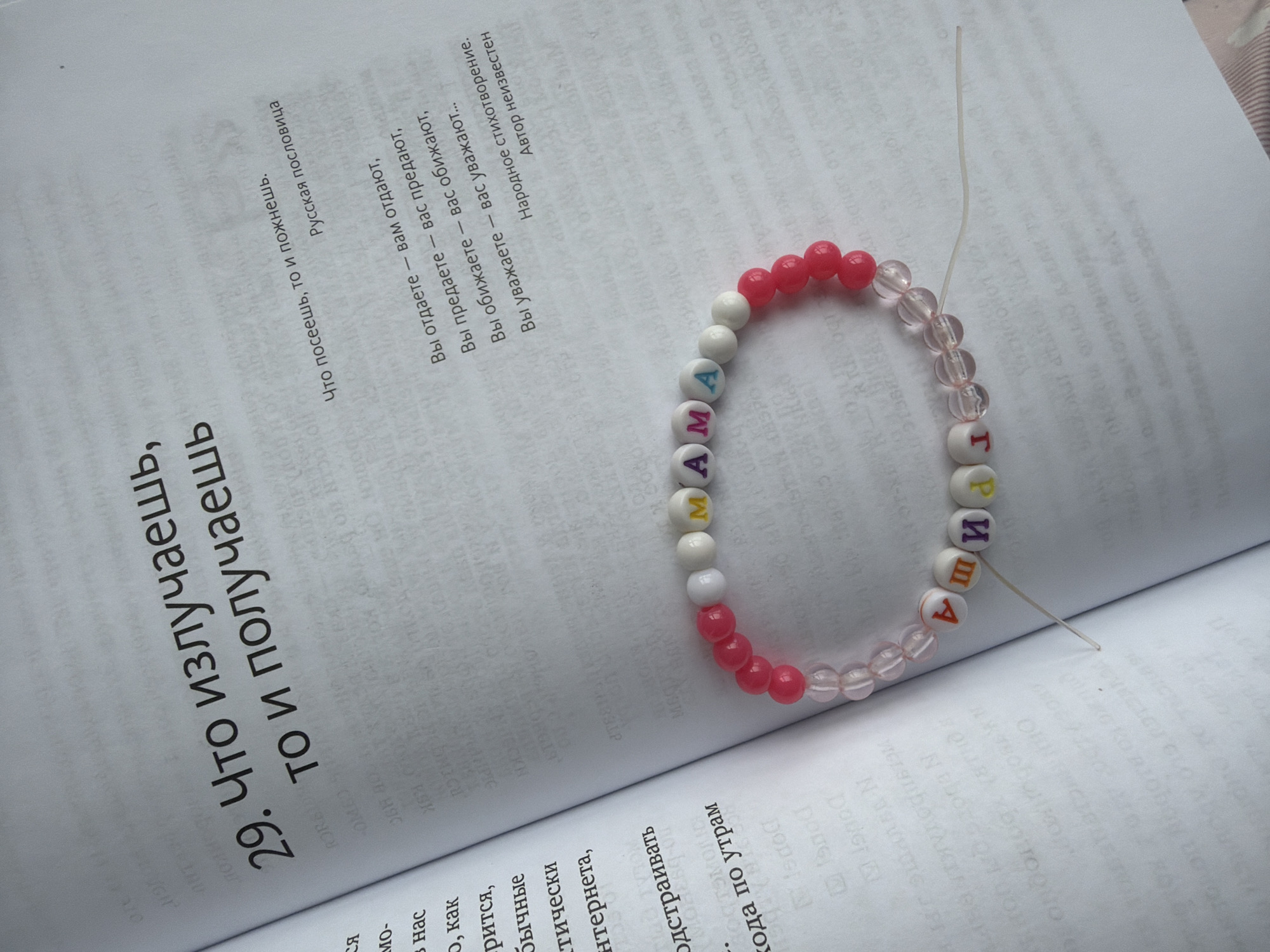 A close-up shot shows an open book with a bracelet laying on its pages. The book is printed in Russian, and the open pages reveal a quote, "29. What you radiate, you receive". The bracelet is made of colorful beads, some pink, some white, and some clear, with lettered beads spelling out a word "MAMA" (mother). The bracelet has not been fully assembled, and a thin, transparent thread runs through the beads. The surrounding environment is the pages of the book, revealing more text and paragraphs in Russian. The location is not explicitly mentioned in the visual information, so it cannot be identified as Izhevsk, Russia.