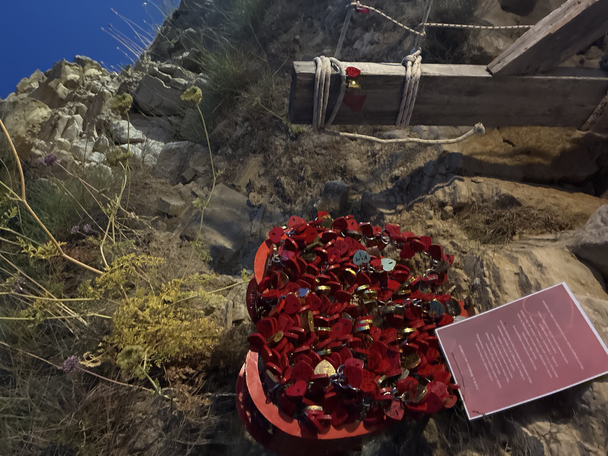 A large, red, heart-shaped sculpture, adorned with numerous small red heart-shaped locks, is mounted on a rocky cliff face. A sign with text is attached to the base of the sculpture. To the right, a wooden post is secured by ropes, with more red heart-shaped locks attached. The environment is natural, with rocks and sparse vegetation under a dark sky. This appears to be a memorial site, possibly located in a coastal area or on a scenic overlook. The location is identified as being in the Laganas Municipal Unit, Greece.