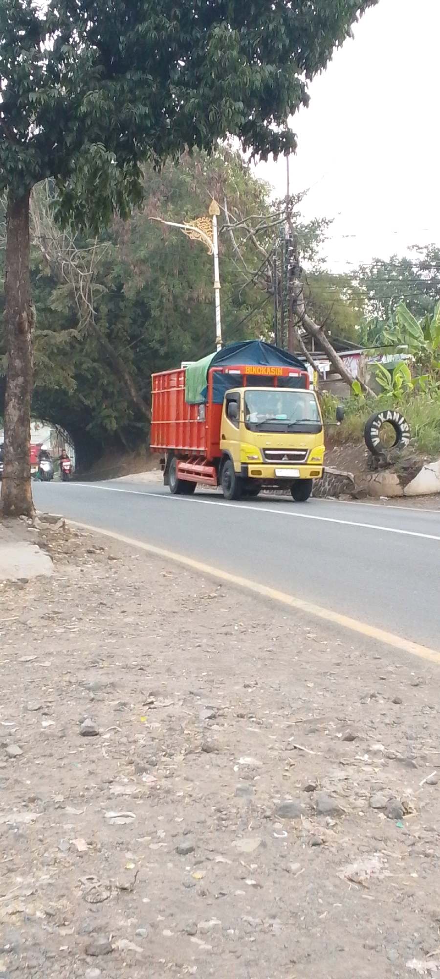 A yellow truck with a red cargo box is driving on a paved road. The truck has a blue tarp covering part of the cargo. The words "BINOKASIH" are visible on the top of the truck's cabin. The road has a yellow line marking the edge. To the left of the road, there is a dirt shoulder with scattered stones and debris. A large tree with dark green leaves is on the far left of the image, casting shadows. Further back, there are more trees and vegetation, and a white street lamp with decorative elements. On the right side of the road, there are more trees, some banana plants, and a round sign with writing. The sky is overcast and white. The image appears to be taken from a low angle, looking up towards the truck. The overall scene suggests a rural or suburban road in a tropical environment. The location is identified as Cilaku, Indonesia, based on external information.