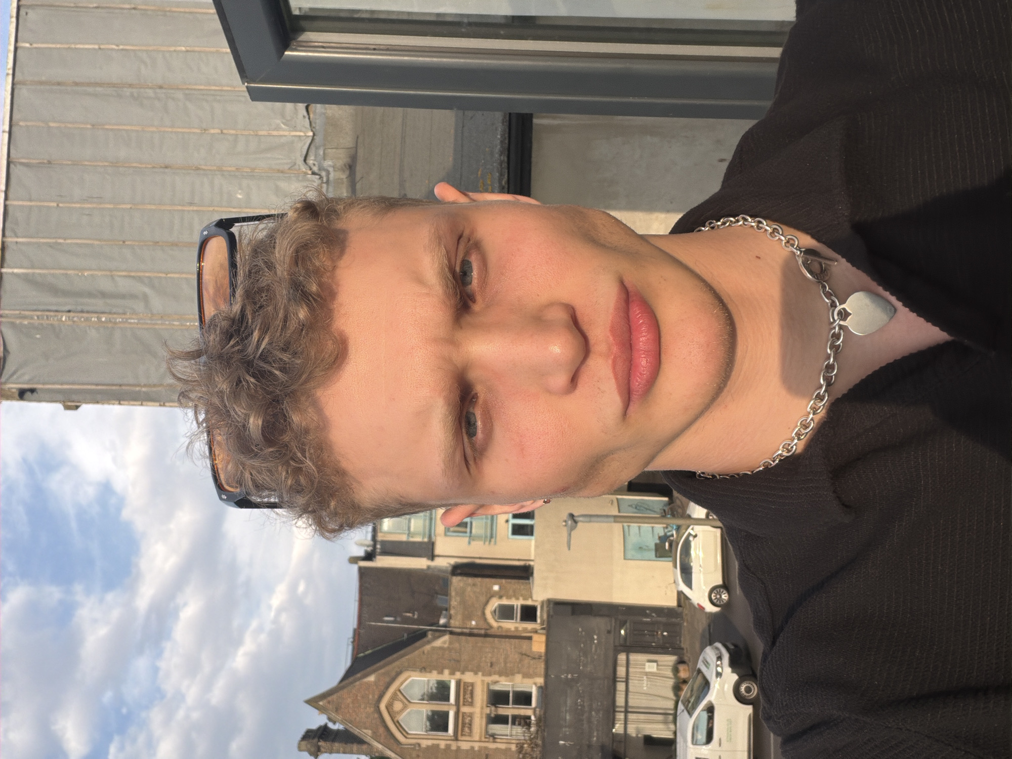 A portrait of a young person with light brown curly hair, fair skin, and light-colored eyes, looking directly at the camera with a neutral expression. They are wearing a dark top and a silver chain necklace featuring a heart-shaped pendant and a toggle clasp. Sunglasses are resting on top of their head. Warm, golden sunlight illuminates the left side of their face, suggesting late afternoon or early evening.
The background depicts an urban street scene in London, United Kingdom. On the left, a bright sky with scattered white clouds is visible. Below the person's right shoulder, there are multiple-story buildings with traditional brick architecture, including prominent arched windows. Several white vans are visible on the street in the mid-ground. Above the person's head, part of a building facade with horizontal siding and a dark window frame can be seen. No legible text is present in the image.