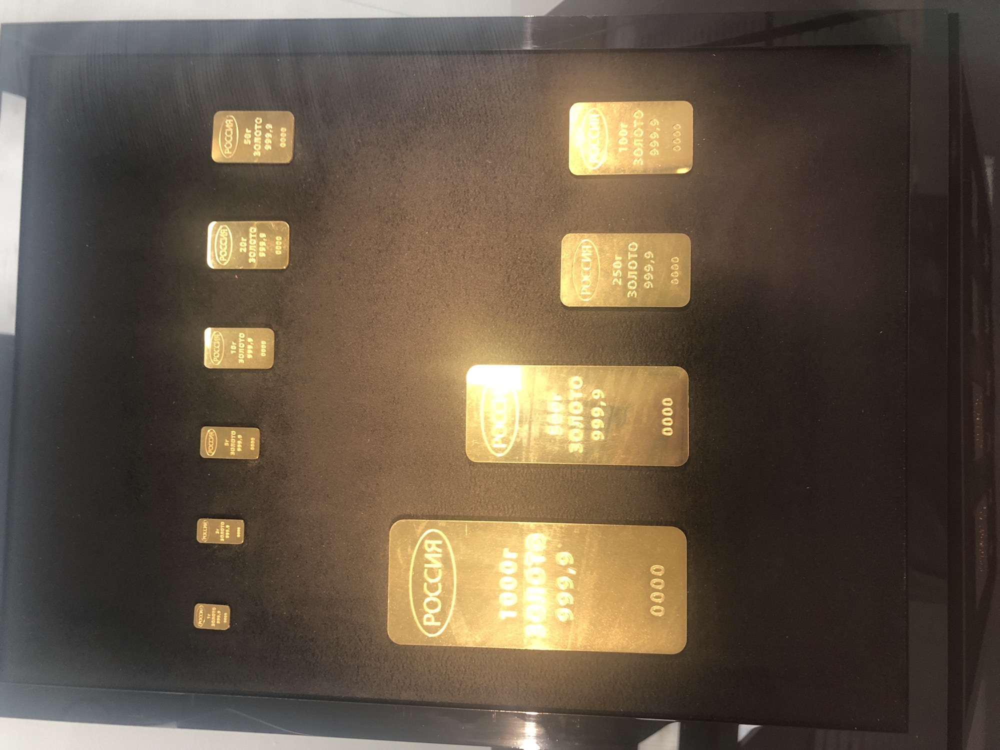 The image displays a collection of gold bars of varying sizes arranged in two rows on a black surface, likely a display case. The top row features six smaller gold bars, while the bottom row contains four larger ones. Each gold bar has a distinctive inscription, primarily in Russian. The larger bars in the bottom row are more prominently featured. The largest bar, on the far left of the bottom row, is inscribed with "РОССИЯ" (Russia) at the top, followed by "1000г ЗОЛОТО 999,9" (1000g Gold 999.9), and "0000" at the bottom. The other bars in the bottom row are also inscribed with "РОССИЯ", followed by their respective weights (500g, 250g, 100g) and the same gold purity (999.9) and serial number format. The smaller bars in the top row appear to have similar inscriptions, though the details are less clear due to their size and distance. The lighting suggests an indoor setting, possibly a museum or a vault display. The overall scene is static and focuses on the gold bars as objects of value and display. There are no people or other discernible objects in the immediate vicinity of the gold bars. The environment is dark and uncluttered, emphasizing the shine and texture of the gold.