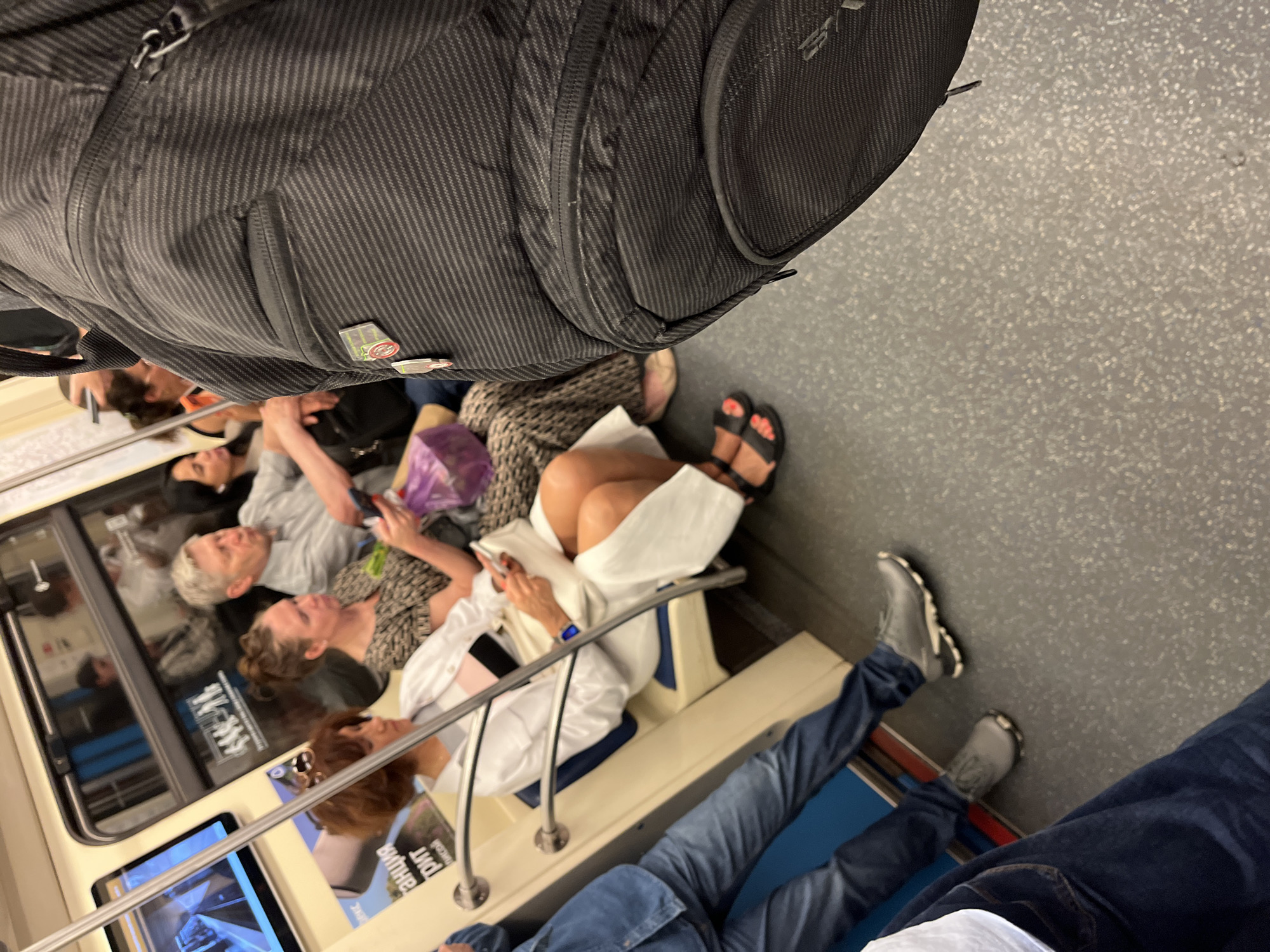 The image shows people seated on a train in Moscow, Russia. The perspective is from above, looking down at the passengers. A large black backpack dominates the top of the frame.
Several individuals are visible, some looking forward and others engaged in their phones. One woman is wearing a white shirt and has her legs crossed, with a white cloth draped over them. Another woman is wearing a patterned dress and black sandals. A man with grey hair is seated, looking to the side.
The train car appears to be relatively full, with passengers seated close together. The floor of the train is a textured grey. Reflections in the windows show more passengers and the interior of the train.
There is some visible text on a poster reflected in the window, but it is not legible. The overall impression is of a crowded commute in an urban environment. The time of day is not specified, but the lighting suggests it is daytime. There are no obvious indications of the weather.