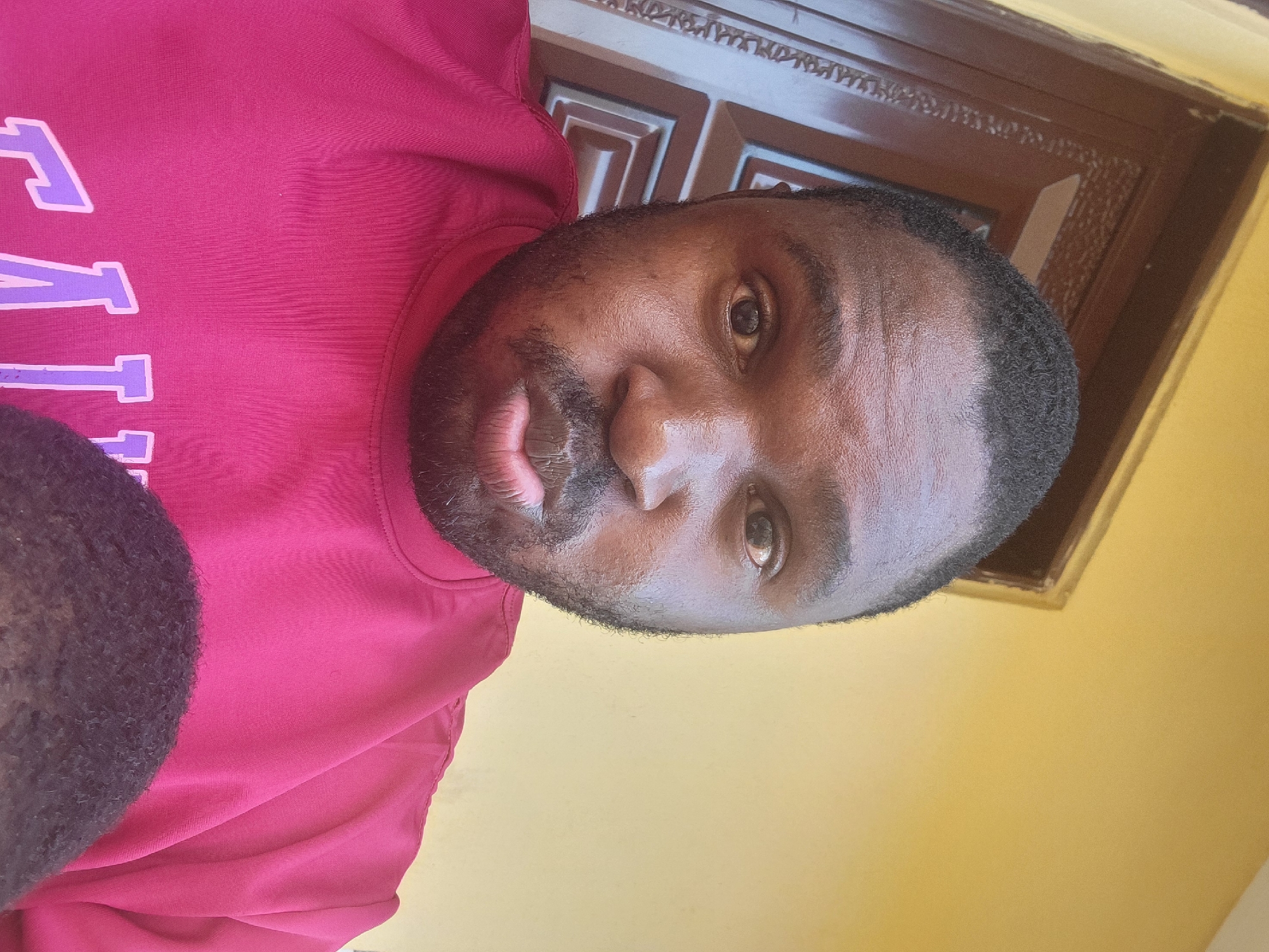 A medium shot depicts a Black man wearing a red shirt with lavender lettering, seemingly a collegiate or team name. He has a trimmed beard and short, dark hair. The background includes a decorated brown door frame and a yellow wall. The location is Suleja, Nigeria.