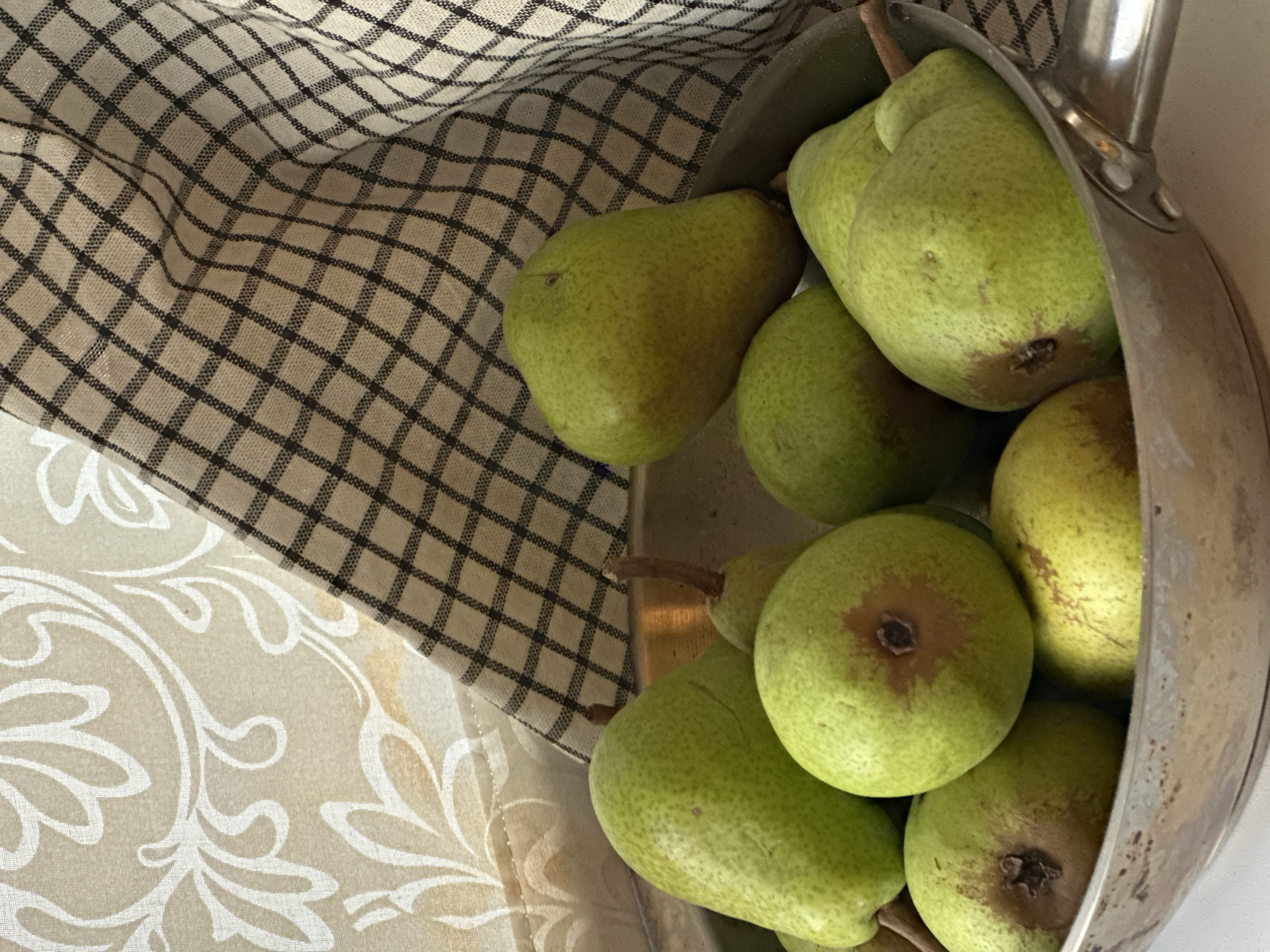 A close-up, slightly overhead shot shows a metal bowl filled with ripe, green pears. The pears are various sizes and shapes, with some showing brown blemishes. Behind the bowl, a light-colored fabric with a white floral pattern is visible on the left, and a checkered cloth with a black and tan pattern is draped on the right. The lighting is soft, casting subtle shadows. The scene appears to be indoors, likely in a kitchen or dining area. There is no indication of the city of Nevinnomyssk, Russia, or any specific environmental context.