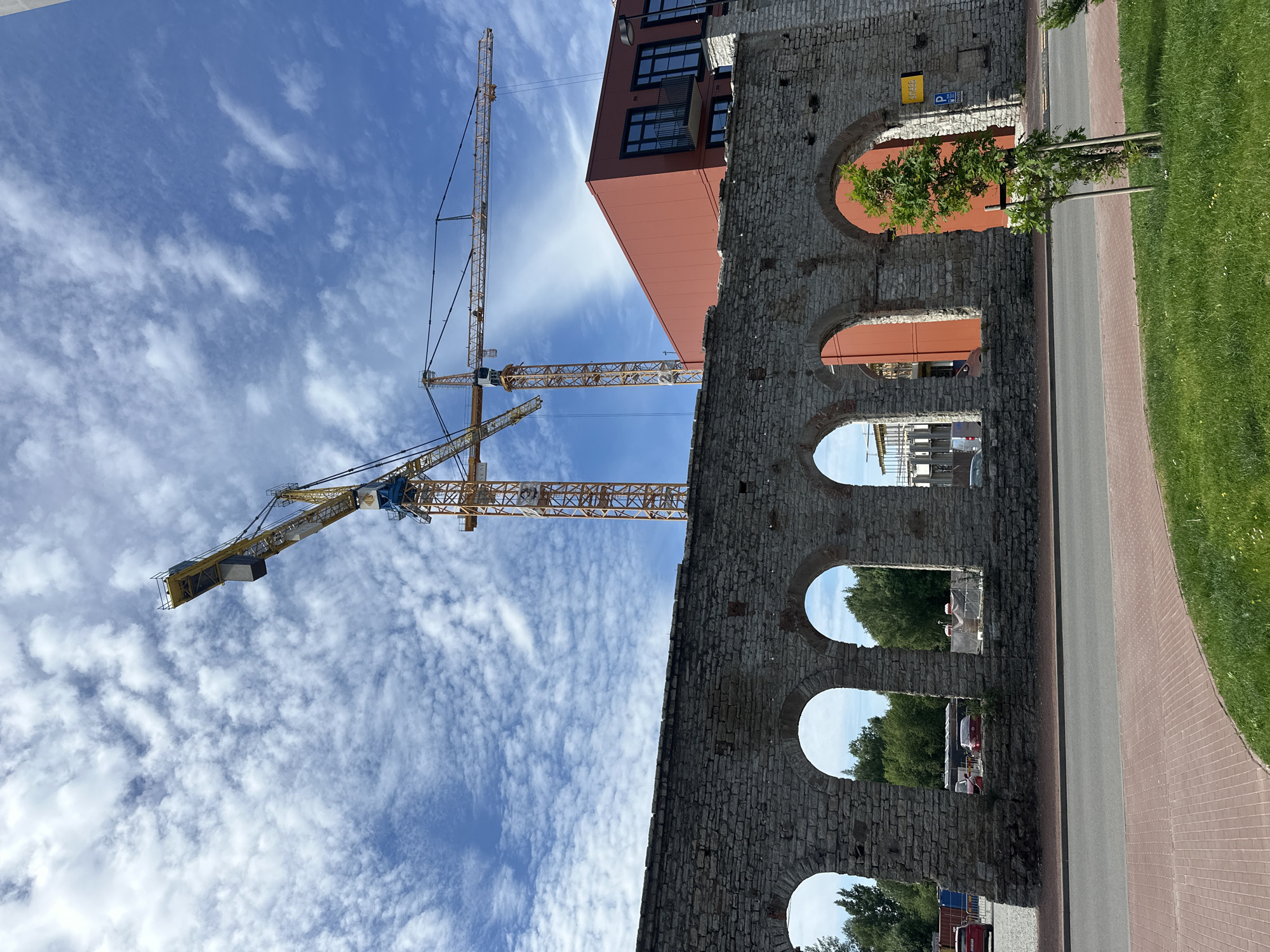 The image depicts a construction site in Tallinn, Estonia, with two yellow tower cranes dominating the sky. In the foreground, a weathered stone wall with several arched openings runs horizontally across the frame. Beyond the wall, a modern red building with dark-framed windows stands tall. The ground in front of the wall features a paved walkway and a grassy area with a young tree. The sky is a vibrant blue, dotted with fluffy white clouds. The scene appears to be a juxtaposition of historical architecture and contemporary development, characteristic of urban renewal projects in European cities.
