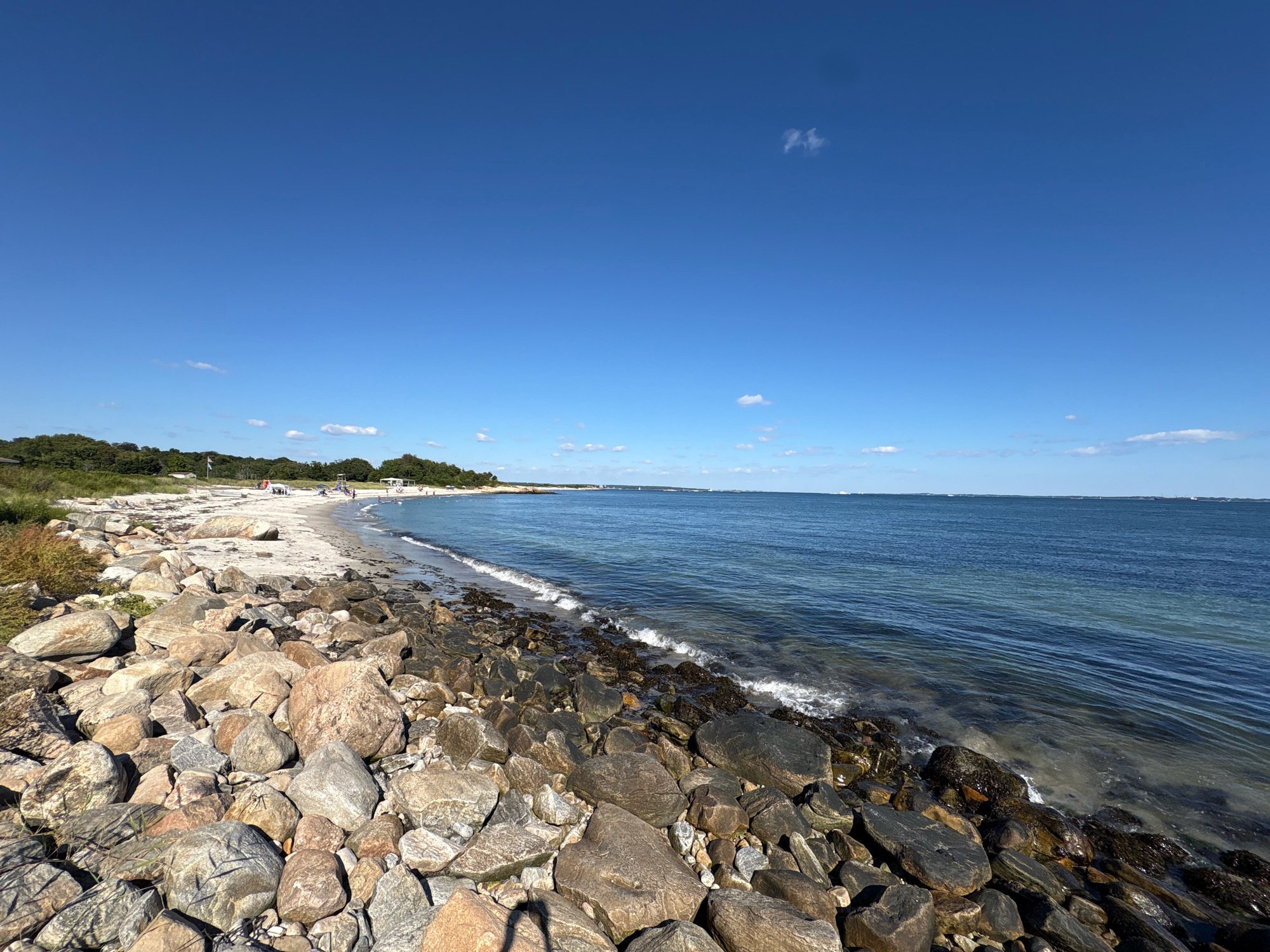 This is a wide-angle, eye-level, outdoor, daytime photograph of a rocky beach and the ocean. The foreground is dominated by a large collection of light brown and gray rocks of various sizes, some of which are wet from the incoming tide. The water is a deep blue, with small waves breaking along the shore and washing over some of the rocks. A sandy beach is visible in the mid-ground, with a line of green trees and vegetation behind it. There are a few scattered people on the beach, some appearing to be sitting or standing. In the far distance, the horizon is marked by a faint line of land and a clear blue sky with a few wispy white clouds. The location is identified as Waterford, United States of America.
