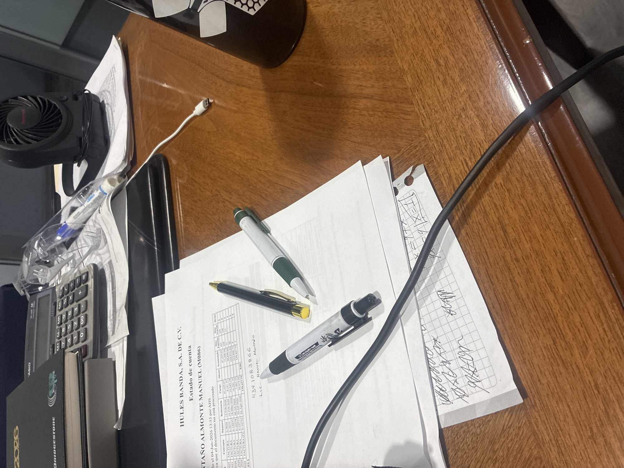 This is a photograph of a wooden desk in an office environment, likely located in Cuautitlán Izcalli, Mexico. The desk is covered with various items, including a black calculator, a stack of papers with financial statements, a black marker, two pens, a small black fan, and a black mug. There are also some handwritten notes on the papers. The overall impression is one of a workspace that is actively being used, with documents and writing instruments laid out. The lighting suggests it is daytime, and the environment appears to be indoors.