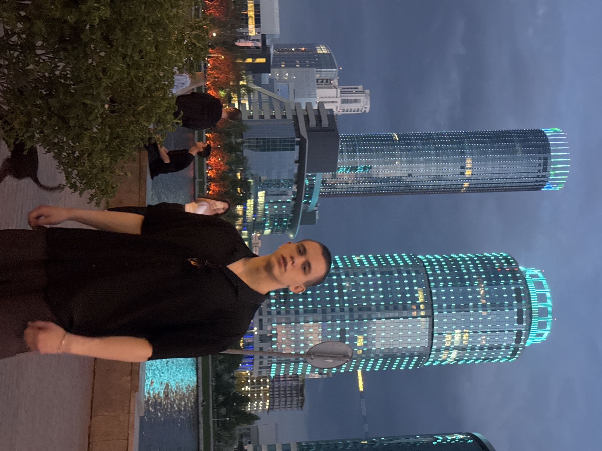 A man stands in the foreground of a nighttime scene in Yekaterinburg, Russia. He faces the camera, wearing a dark shirt and pants. Behind him is the Iset River. Across the river, buildings are illuminated with blue and white lights, including the cylindrical Vysotsky skyscraper and a second skyscraper. Other individuals are visible, including someone sitting on a ledge and several people walking. A bush partially obscures the left side of the scene.
