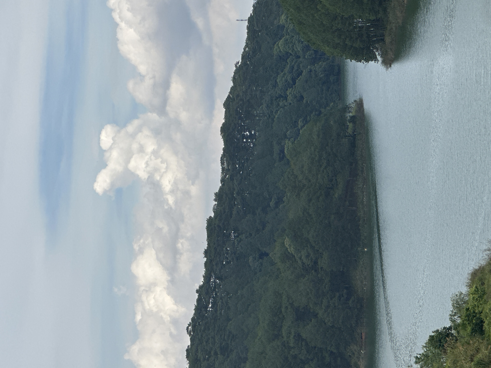 The image depicts a landscape of a lake surrounded by lush, green, forested hills. The water appears calm with gentle ripples on its surface. The sky is filled with a mix of blue patches and puffy white clouds. A cellular tower is present on the top of the mountain. The location is likely in Da Lat, Vietnam.