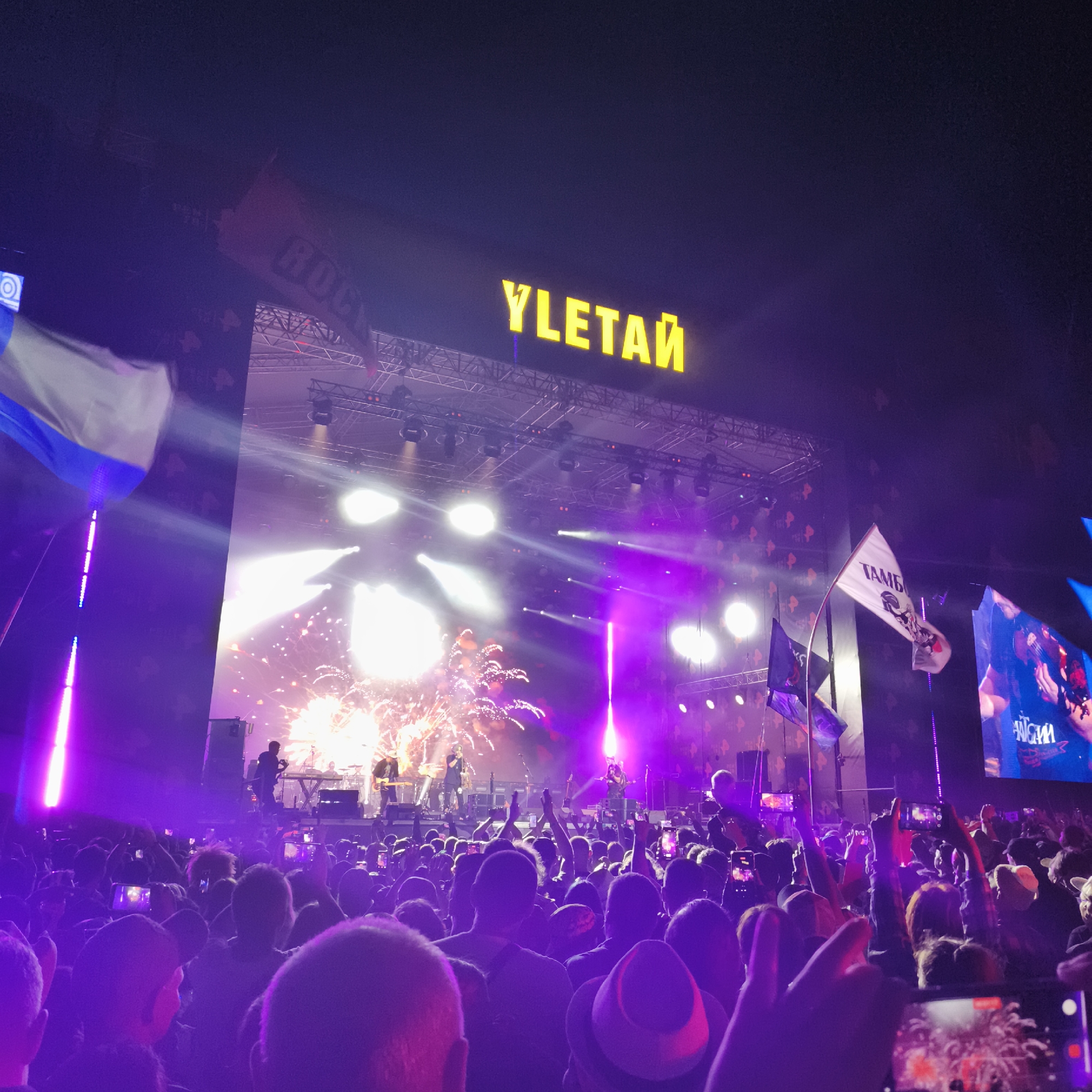 This media file depicts a lively outdoor concert scene at night, likely in Lagunovo, Russia, given the context. A large crowd of people are gathered, illuminated by vibrant purple stage lighting and the bright glow of fireworks exploding above the stage. Many in the audience are holding up their phones, capturing the spectacle.
The stage is set up with multiple lighting rigs and speakers, and a band is performing. The main focal point of the stage is a large, illuminated sign spelling out "YLETAй" in bright yellow capital letters. There are also flags visible, one white flag with text and an illustration, and a dark blue flag, both held aloft by poles. The overall atmosphere appears energetic and celebratory, with the crowd seemingly engrossed in the performance. The time of day is clearly night, suggested by the dark sky and the dominance of artificial lighting. The weather is not discernible from the image. The text "YLETAй" is prominently displayed on the stage sign. The white flag has the text "ТАМБ" on it.