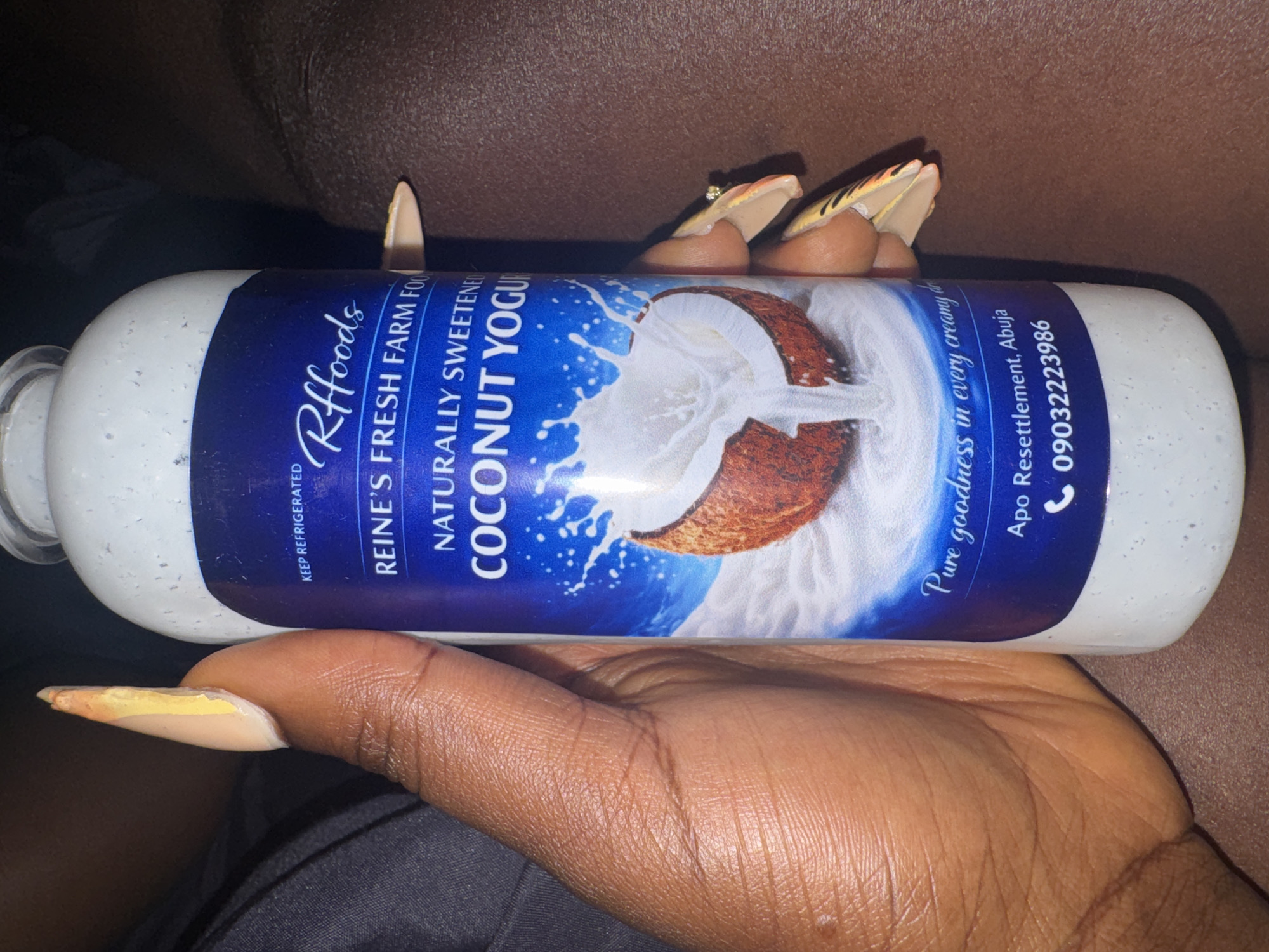 A person's hand with long, decorated nails holds a bottle of "Rfooods Naturally Sweetened Coconut Yogurt." The label is blue with white text and features an image of a coconut with milk splashing out. Below the image, text reads "Pure goodness in every creamy d," followed by "Apo Resettlement, Abuja" and a phone number "09032223986." The background is dark and out of focus, suggesting an indoor setting. The hand appears to be resting on a textured surface.