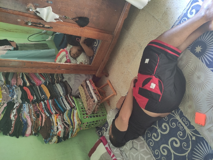 The image shows a man lying on a bed in a room in Kecamatan Lawang, Indonesia, located at latitude -7.828 and longitude 112.701. The man is wearing black shorts with a red stripe and a black shirt. His right leg is bent at the knee and resting on the bed. The bed has a blue and white patterned cover. In the background, there is an open wooden cabinet with many clothes stacked on the shelves. A green plastic basket is on the floor, with a patterned fabric behind it. There is also a small, orange object on the bed next to the man. The room has a green wall and a white tiled floor. The man's right hand is hanging off the bed, and his left hand is resting on the bed. He appears to be resting.