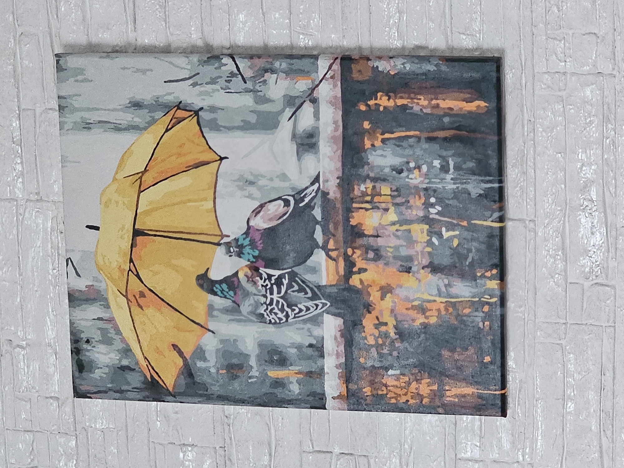 The image shows a painting of two pigeons huddled together under a yellow umbrella. The painting is hanging on a white brick wall. The pigeons are gray and have iridescent patches of blue and purple on their necks. They are perched on a ledge, and the background appears to be a rainy city scene with blurred lights. The bottom of the painting depicts reflections of lights on a wet surface, possibly a street or pavement. The overall style of the painting is impressionistic. The location is not specified, and there is no indication of Kalinovka, Russia.