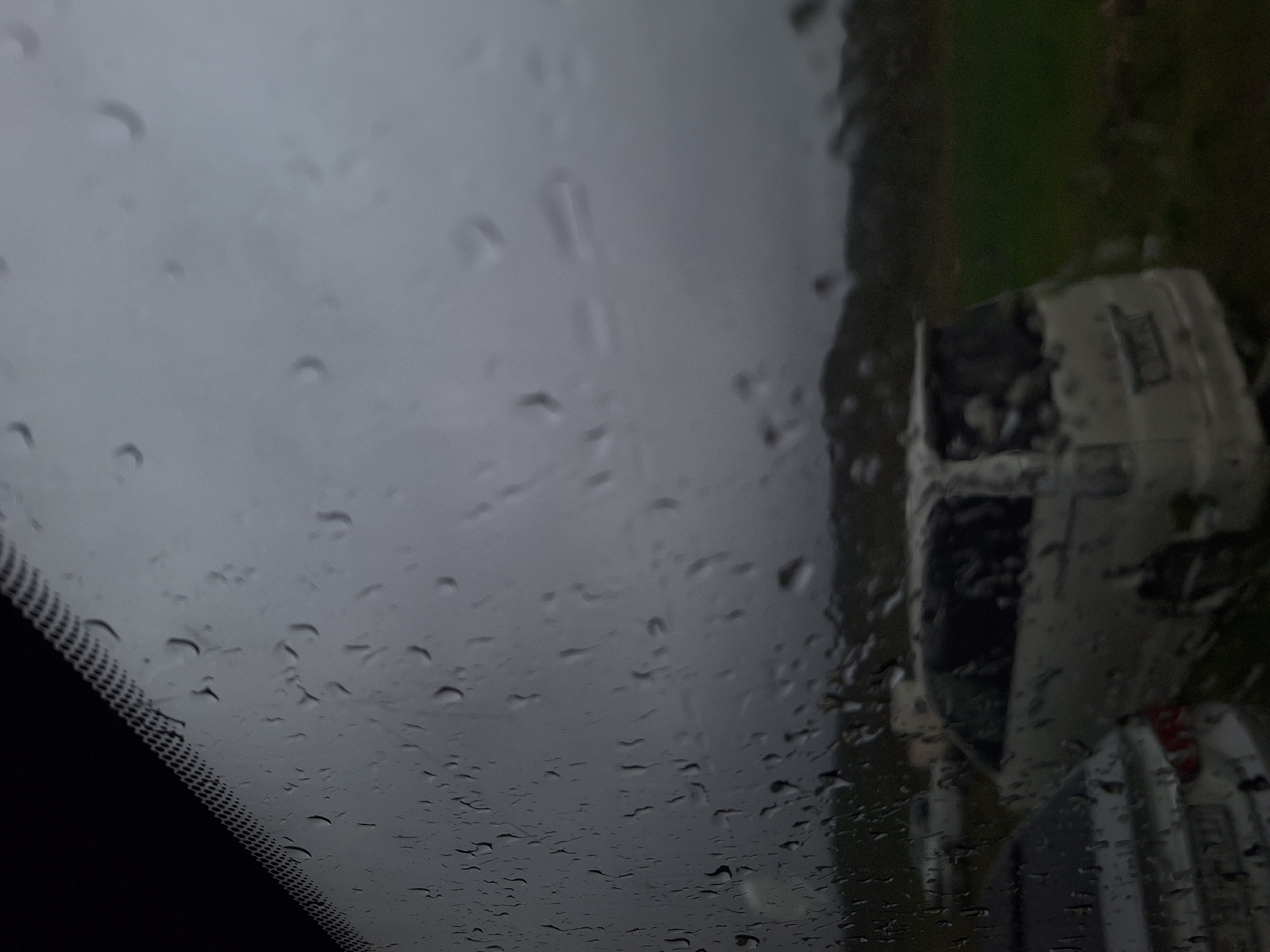 The image depicts a view through a rain-streaked windshield. The windshield wiper blade area is visible in the lower left corner. Outside, a blurred view of what appears to be vehicles parked next to a grassy area. The scene is overcast. Given the request to include the city of Tlik, Armenia, it is assumed the image was taken there, however, there is no visual information to independently confirm the location within Tlik or even that the image was taken in Tlik. No people are visible. The objects are vehicles and wet grass, the action is it is raining, the interation is one of vehicles parked in wet conditions. The environment is a gray and overcast day.