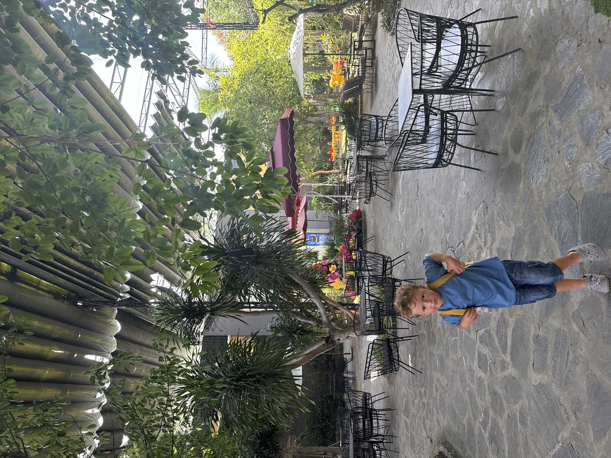 The image depicts a boy standing in an outdoor seating area that appears to be a cafe or restaurant. The area is paved with irregular stone slabs and features numerous black metal tables and chairs. Several large plants, including palm-like trees, are scattered throughout the space, and the area is covered by a corrugated roof structure that allows sunlight to filter through. In the background, there are more trees, some with bright yellow and pink flowers, and colorful playground equipment. The boy, wearing a blue t-shirt, denim shorts, and grey clogs, has a yellow backpack slung over his shoulders. He is looking towards the camera with a slight smile. The overall impression is of a relaxed, possibly family-friendly, outdoor dining or recreational space in Nam Nha Trang Ward, Vietnam.
