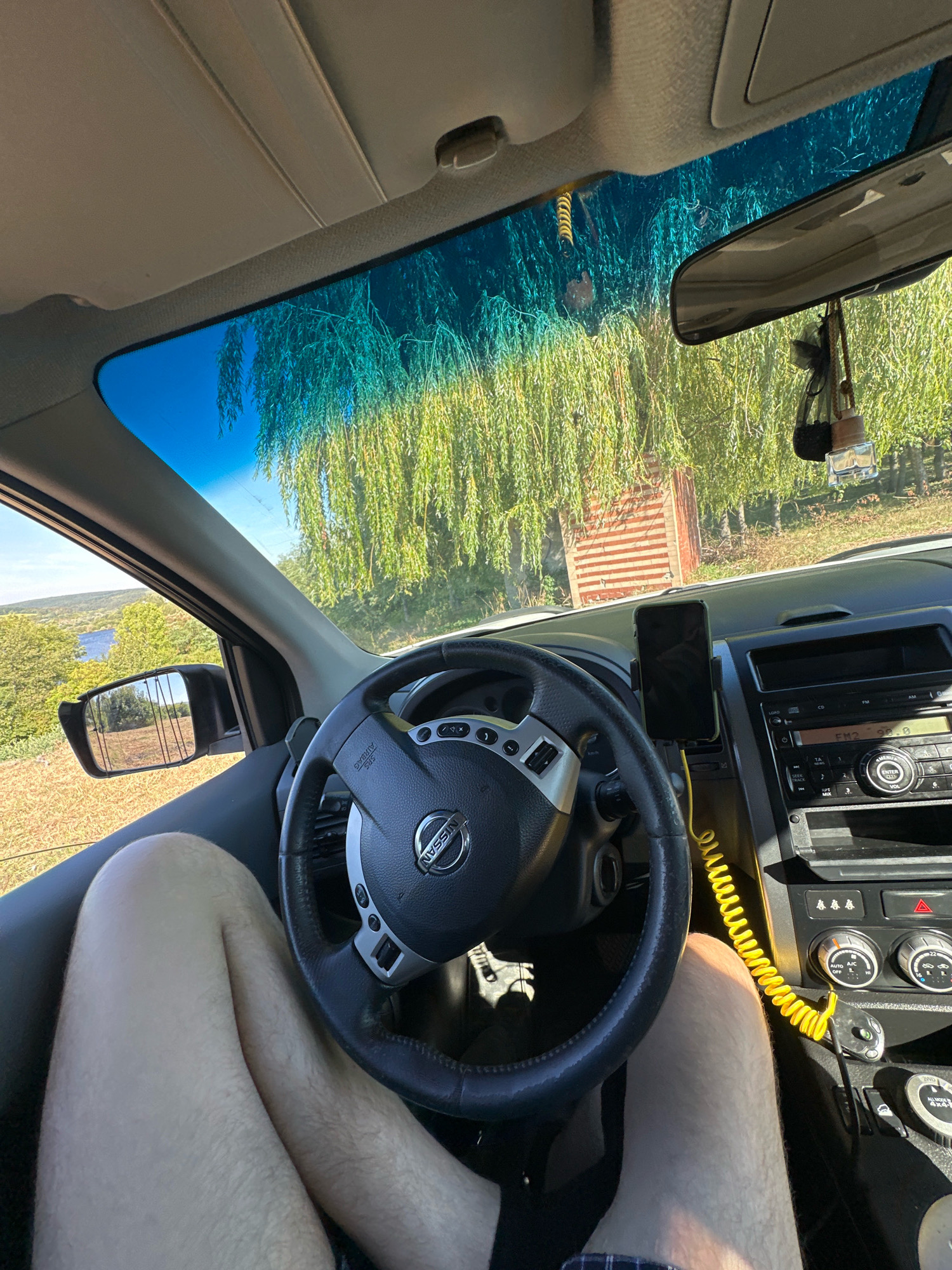 The image provides an interior view from the driver's seat of a car, looking forward through the windshield and out the side window. The driver's legs are visible in the foreground, positioned in front of the steering wheel. The steering wheel, dashboard, and center console of the car are prominent features. A smartphone is mounted on the dashboard to the right of the steering wheel. Outside the car, through the windshield, a weeping willow tree with dense foliage dominates the view, with a glimpse of a blue body of water and rolling hills in the distance to the left. A rustic fence or structure is partially visible behind the tree. The overall lighting suggests it is daytime. No specific landmarks or indicators point to the exact location within Makiv, Ukraine, beyond the general context of a rural or semi-rural setting.