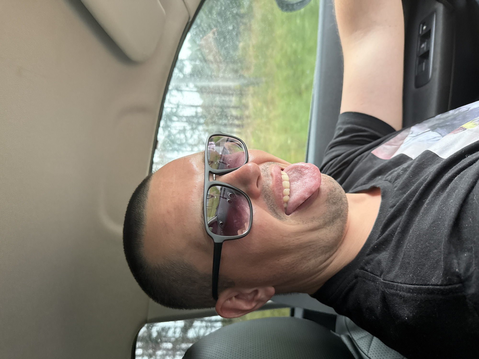 A man with a buzz cut and sunglasses is sticking his tongue out while driving in a car. He is wearing a black t-shirt. The scene appears to be in a rural area outside of a city, as suggested by the trees and green fields visible through the car window. It is not possible to determine the specific location within Podshivalovo, Russia from the image.