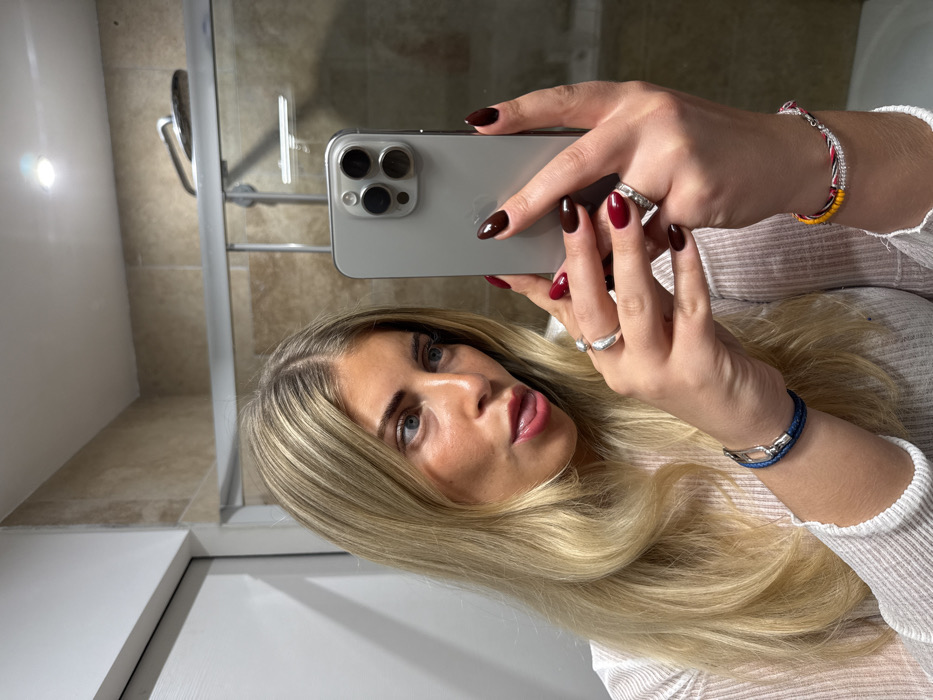 A woman holds a smartphone up to take a selfie. She has light blonde hair, fair skin, and blue eyes. She is wearing rings and bracelets. Her nails are painted with a deep, dark red polish. The background appears to be a bathroom, with a shower and tiled walls visible in the mirror's reflection.